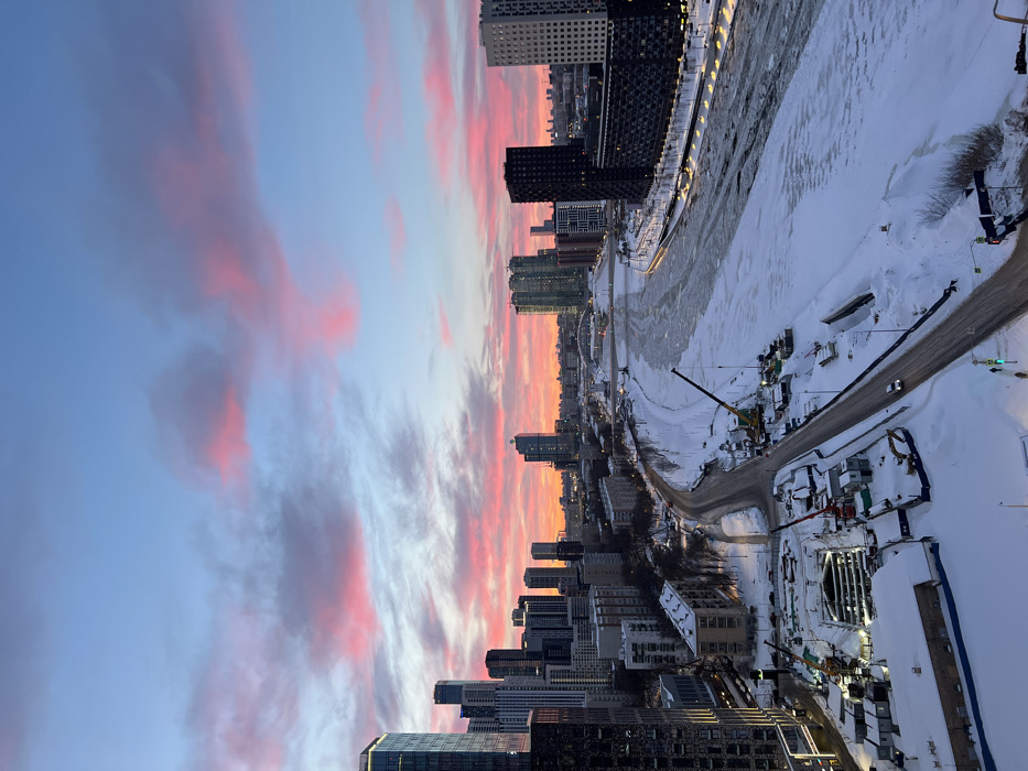 A vibrant cityscape at dusk features a pink and orange sky reflected in scattered clouds. Below, a river runs alongside modern buildings, some under construction. A road, blanketed with snow, winds along the river's edge. Construction equipment and vehicles are visible, adding to the scene's urban energy. The image captures the contrast between natural beauty and urban development.
