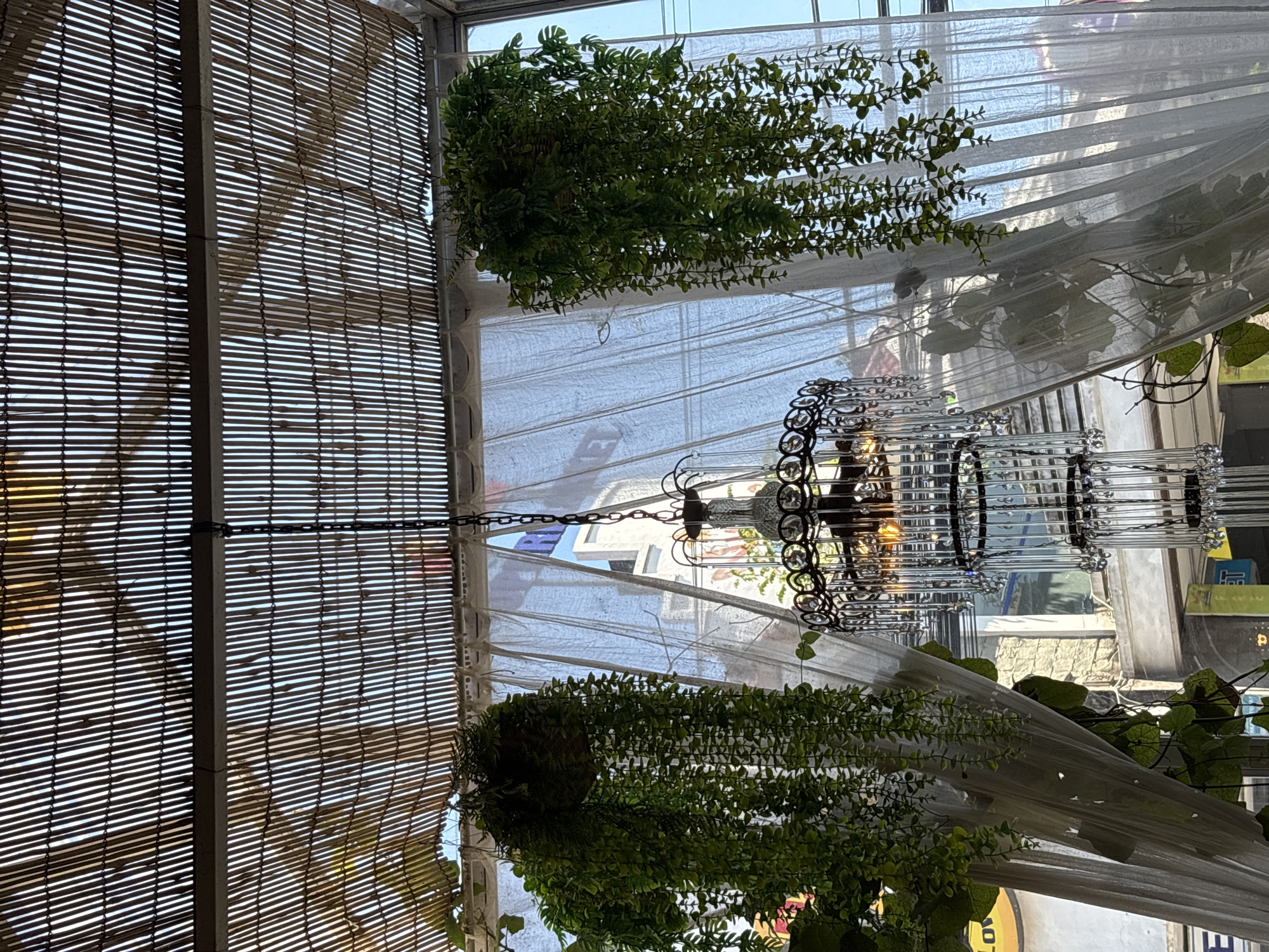 The image captures an interior view in Kuta Selatan, Indonesia, featuring a textured bamboo roof section at the left. Greenery hangs from the ceiling, interspersed with sheer white fabric. A multi-tiered metal chandelier hangs from a chain. Through the translucent fabric, buildings and signage are visible.