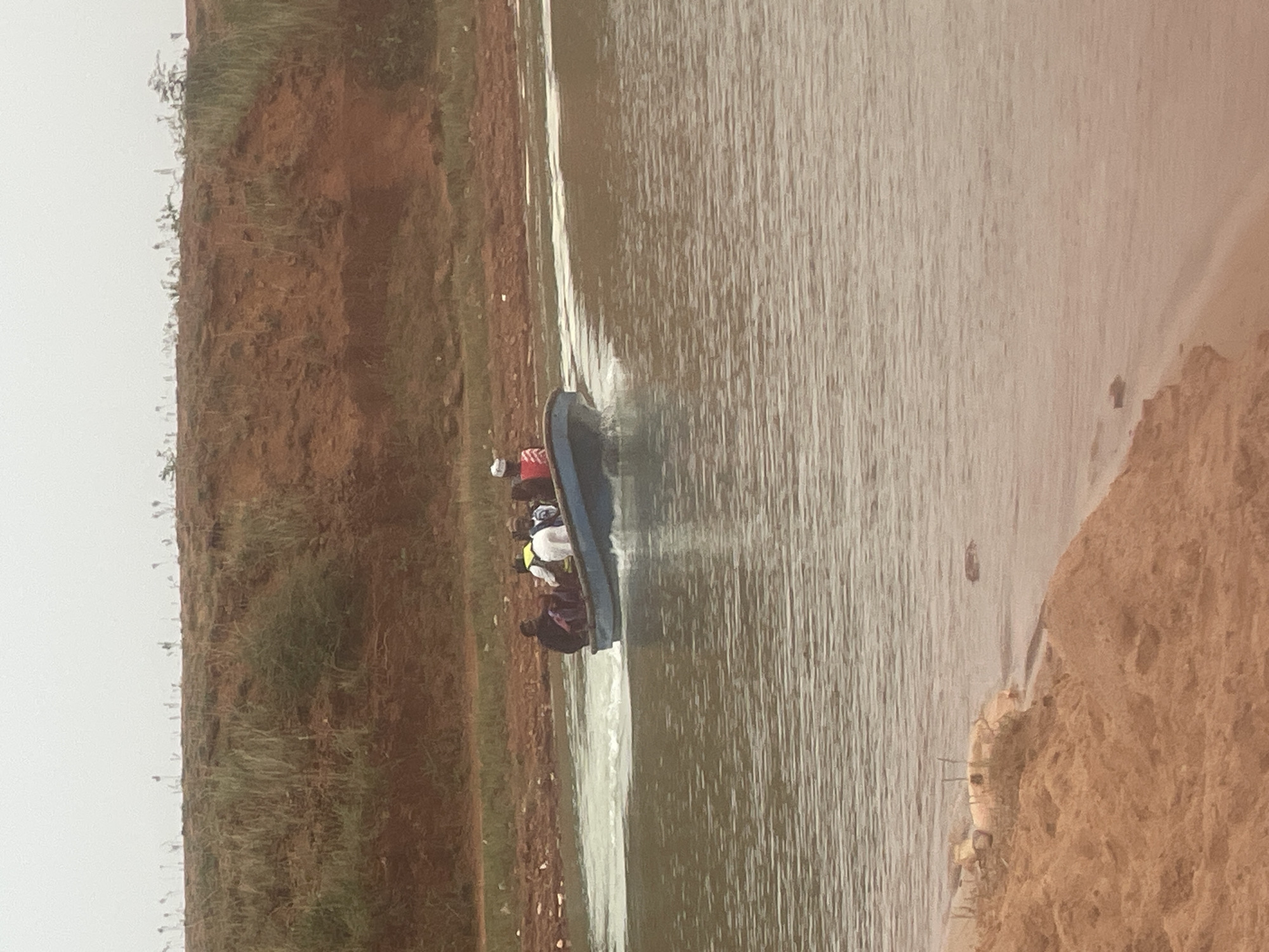 A boat with several people is traveling on a body of water bordered by red soil. The boat is creating a wake. The location is Jos, Nigeria.