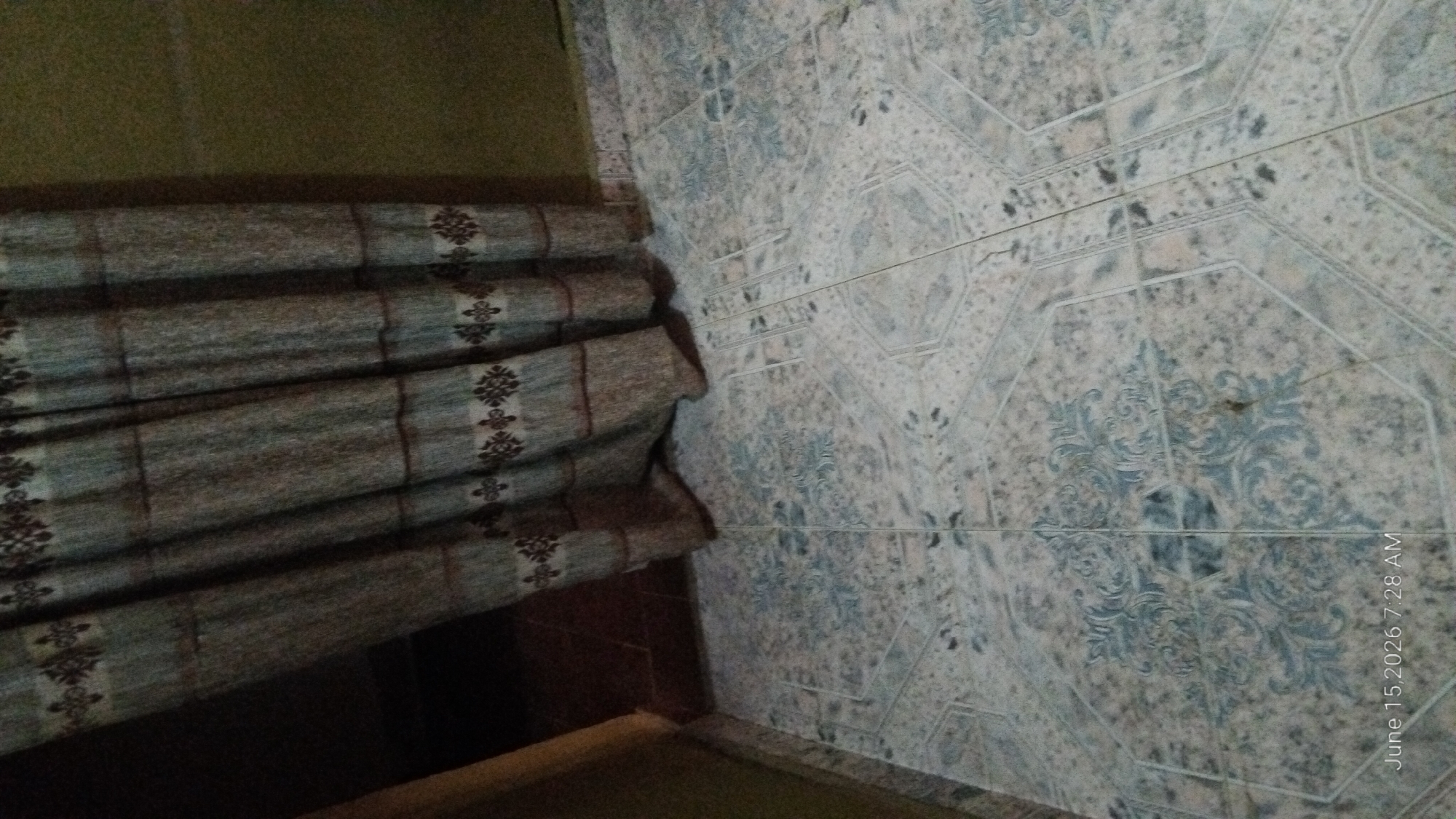 The image shows a tiled floor and a curtain. The floor tiles are light-colored with a blue patterned design. The curtain is a muted green and brown striped fabric with a repeating floral motif. The lighting suggests an indoor environment. There is text at the bottom of the image indicating "June 15, 2026 7:28 AM". This appears to be a timestamp from a camera or device, not an indication of the actual location or time. There is no visible information or context to suggest this scene is located in Howayi, Nigeria.