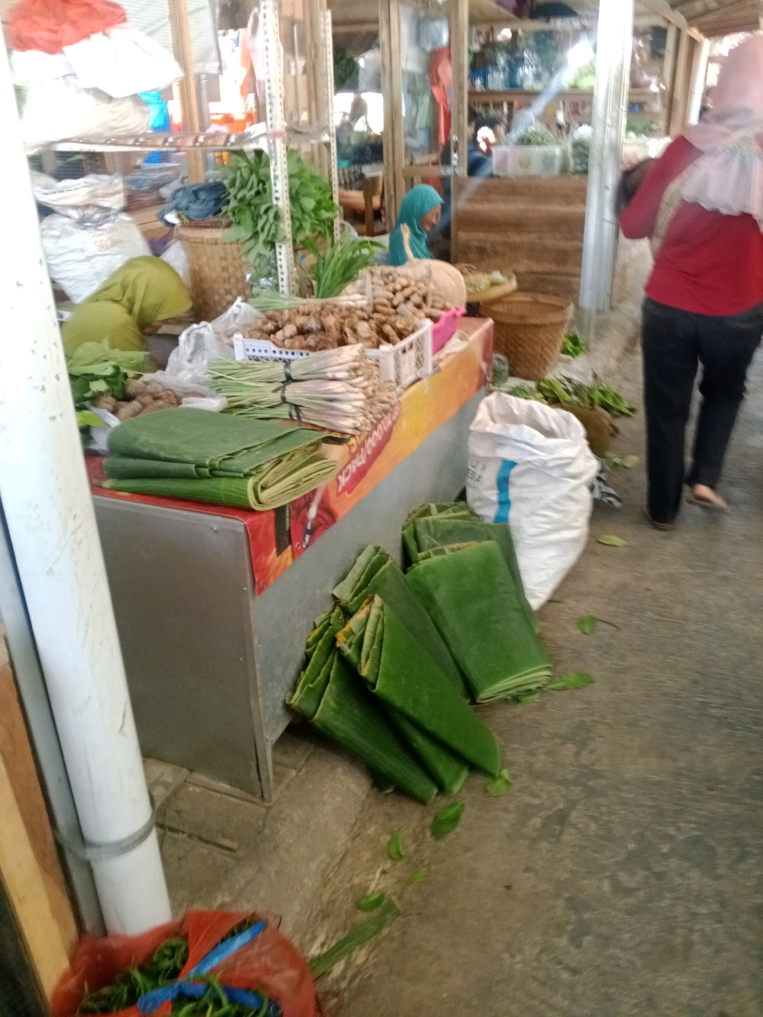 The image depicts a market scene in Wonogiri, Indonesia. In the foreground, folded banana leaves are piled on a gray surface, with more leaves scattered on the ground. To the left, a large white pipe is visible, secured with a gray tie. A red plastic bag filled with green produce, possibly chilies or beans, lies at the bottom left of the frame. In the mid-ground, a stall displays various goods. Folded green leaves are neatly stacked on a counter, along with bundles of ginger and lemongrass. A woman wearing a blue hijab is seated behind the stall, seemingly interacting with the produce. Behind her, shelves are stocked with various items, including large sacks and baskets. In the background, a woman in a red top and pink hijab walks away from the camera. The overall environment appears to be an outdoor or semi-enclosed market with wooden structures and a concrete floor.