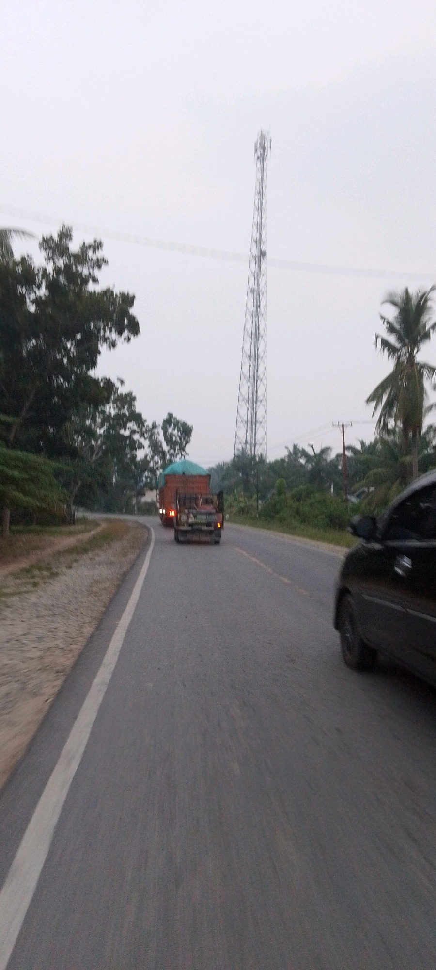 A high-angle, wide shot captures a twilight scene on a paved road in Teluk Pulau Hulu, Indonesia. The sky is overcast and a muted gray, with a tall, lattice-like tower, possibly a communication tower, dominating the mid-ground on the right. To the left of the road, dense green foliage lines the shoulder, obscuring the terrain beyond. A black car is partially visible in the foreground on the right side of the road, its side and rear window in view. Further down the road, a flatbed truck with a green tarp covering its cargo is traveling away from the camera, with a smaller truck or utility vehicle behind it. The road itself is gray asphalt with a faint yellow line marking the center. The motion blur indicates the photograph was taken from a moving vehicle.