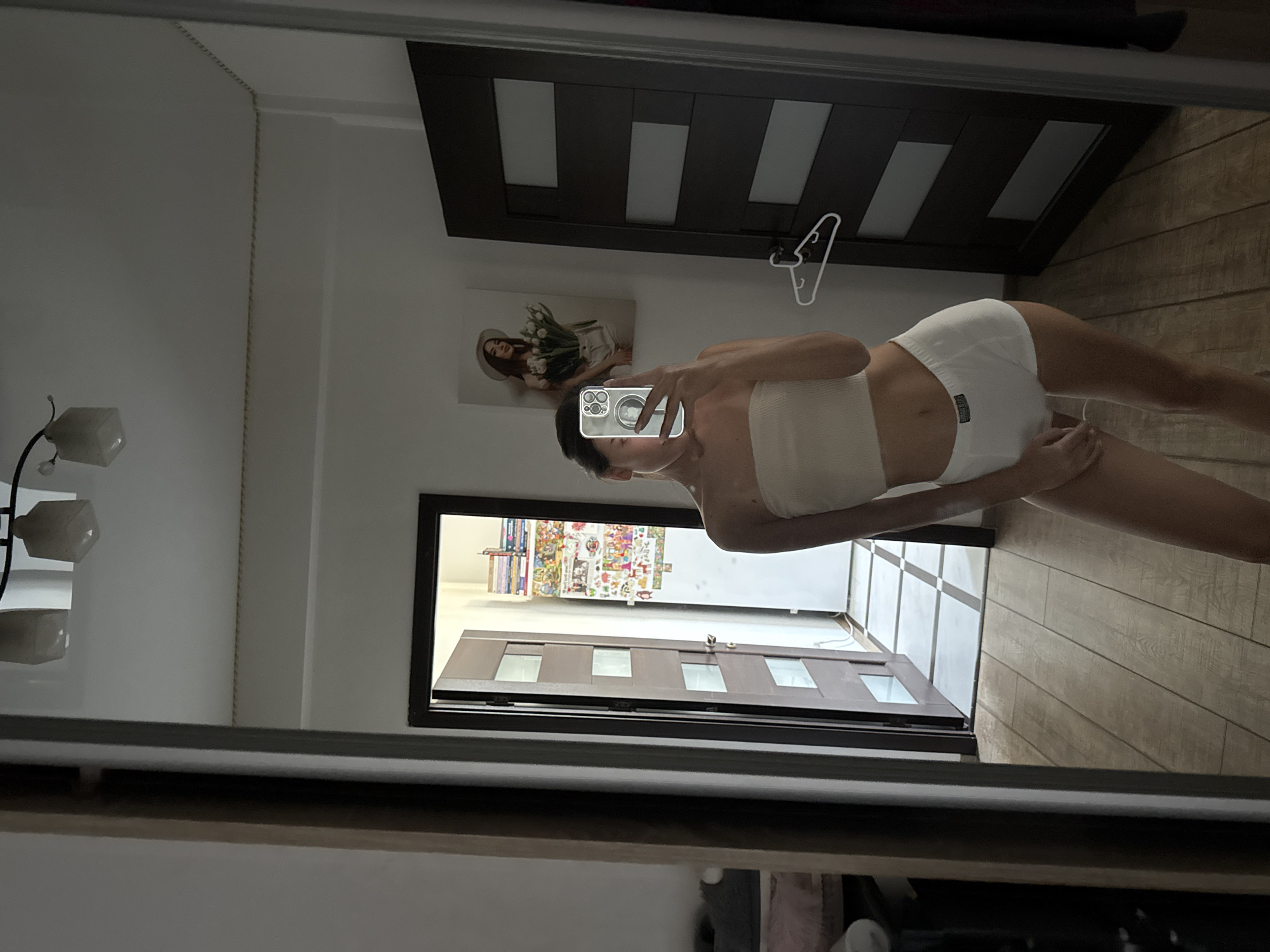 A woman stands in front of a mirror, taking a selfie. She is wearing a white bandeau top and white boxer shorts. Her reflection shows her holding a smartphone with a clear case. Behind her, a doorway leads to another room, where a refrigerator is covered in stickers and magnets. To the right of the doorway, a dark wooden door is visible. The room has wooden flooring and a white ceiling with a modern light fixture. A framed picture of a woman holding flowers hangs on the wall. The scene is set in what appears to be a residential interior, likely an apartment or house, potentially located in Chișinău, Moldova, given the context provided.