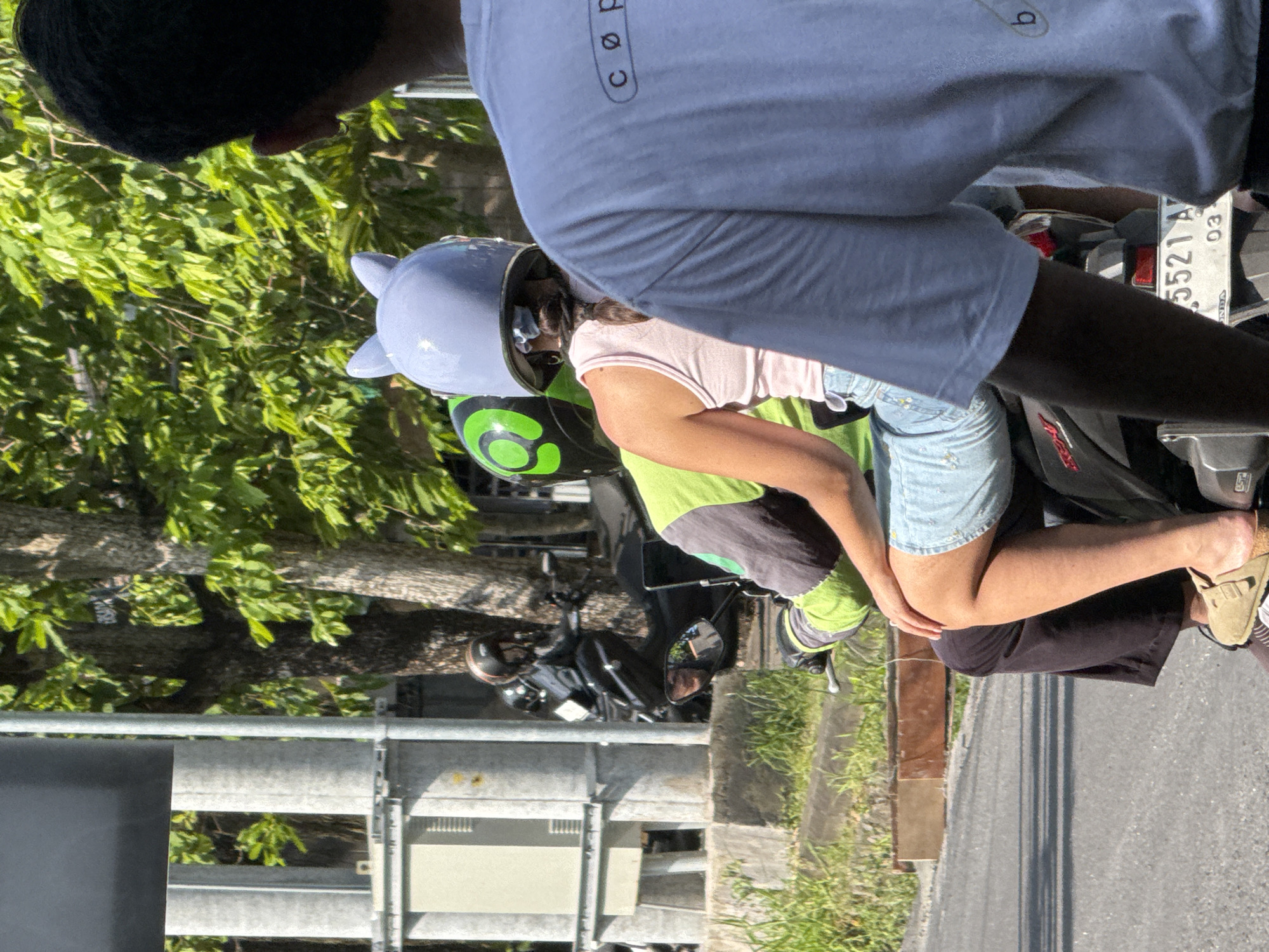 A sunlit street in North Kuta, Indonesia, is depicted with three people on motorcycles. A passenger wearing a light blue helmet with ears is sitting behind a driver in a green-accented jacket. Another person wearing a white shirt stands nearby. Further down the road, another individual rides a separate motorcycle. A metallic guardrail lines the road, and trees are in the background.