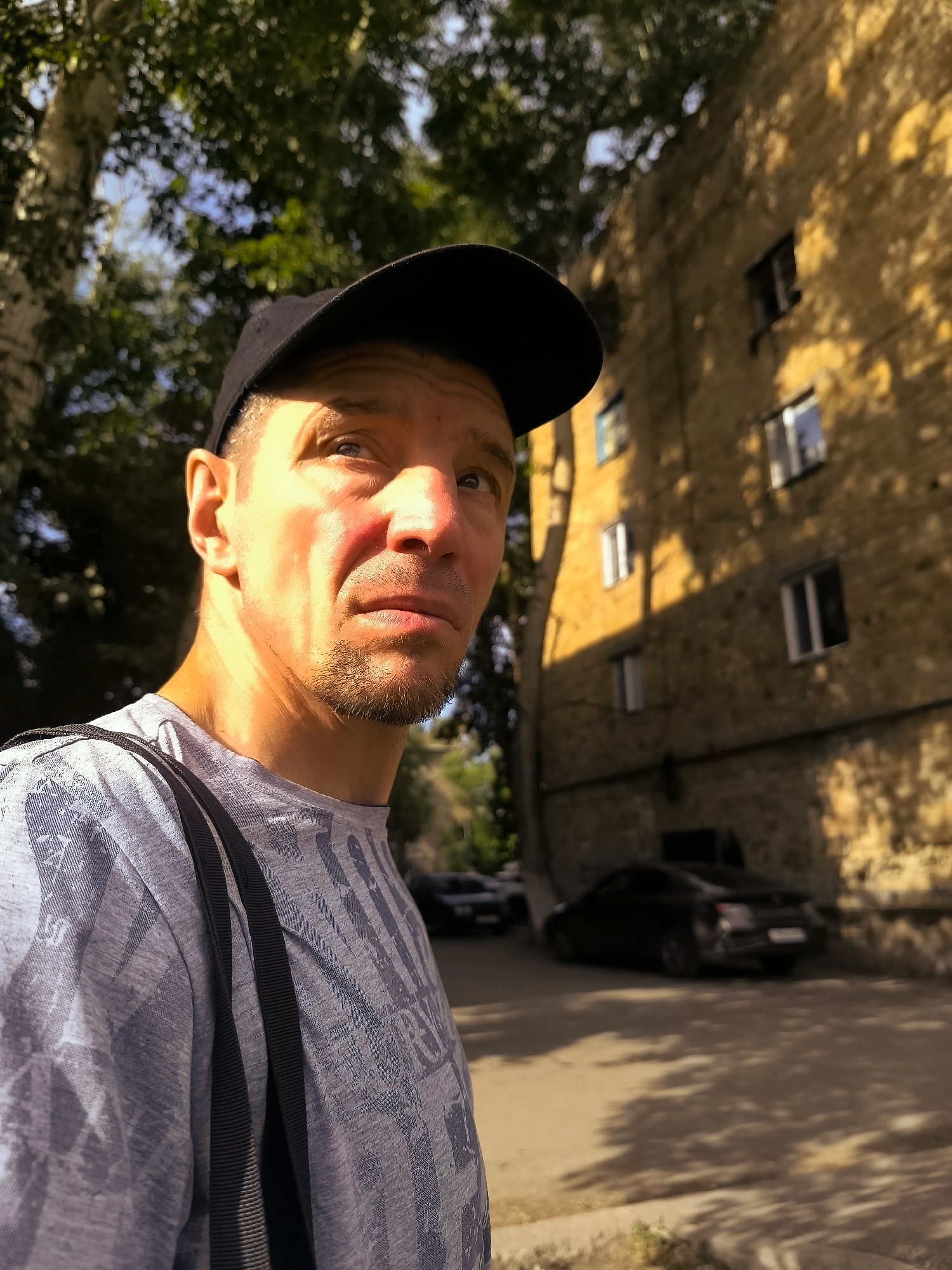 A man wearing a black baseball cap and a light grey t-shirt with a patterned design stands in the foreground, looking to his left. His face is illuminated by harsh sunlight, casting shadows and highlighting his features. Behind him, a yellow brick building with multiple windows is visible, with trees and their dappled shadows. A black car is parked on the street in front of the building. The scene appears to be an urban environment, possibly in Qarağandı city administration, Kazakhstan, given the lighting and architecture.