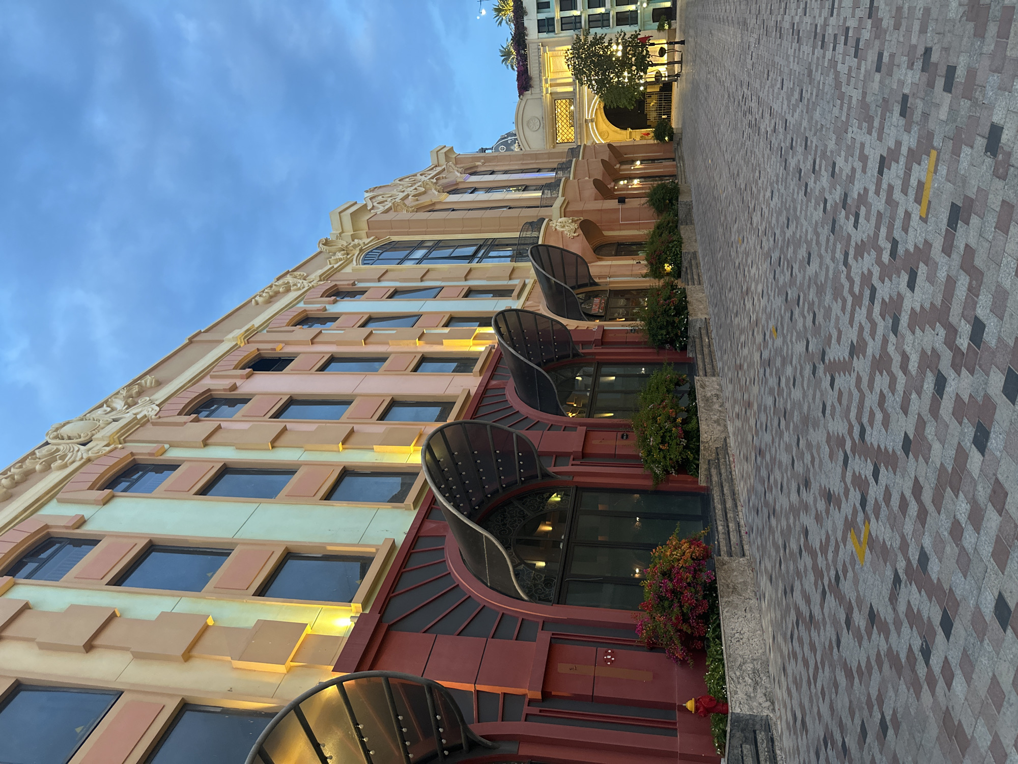 The image captures a street scene in an unknown city. The main subject is a multi-story building with a distinctive architectural style. The building's facade features a mix of colors, including beige, pink, and a light green, with rectangular windows across multiple floors. Some windows have arched, metal-framed projections extending outwards. Lush greenery and flowers are planted in containers along the base of the building. The street is paved with a patterned combination of gray, black, and red bricks or stones. A blue sky is visible above the building. The perspective is from a slightly low angle, emphasizing the height of the building. In the background, another building is partially visible, illuminated with warm lighting. The scene appears to be taken during the evening.