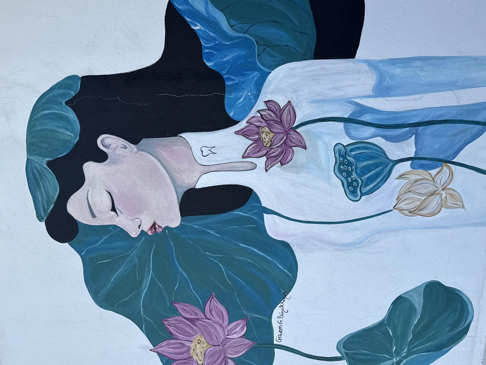 This is a mural on a white wall in Silifke, Turkey. The mural depicts a woman with her eyes closed, shown in profile, facing left. She has dark hair and is wearing a white traditional Vietnamese dress called an "áo dài." The dress has a high collar with a small, stylized tooth symbol on the right side. Around the woman are large, stylized lotus flowers and leaves in shades of pink, purple, teal, and blue. One pink lotus is positioned near her left shoulder, and another is on her chest. A teal lotus seed pod is also visible on her chest, and a beige lotus hangs lower on the right side. Large teal and blue leaves frame the woman's head and shoulders. Below the lotus on the left shoulder, the artist's signature "Gizem G. Büyükkaya" is visible in cursive. The mural has a slightly textured appearance, characteristic of paint applied to a stucco wall.
