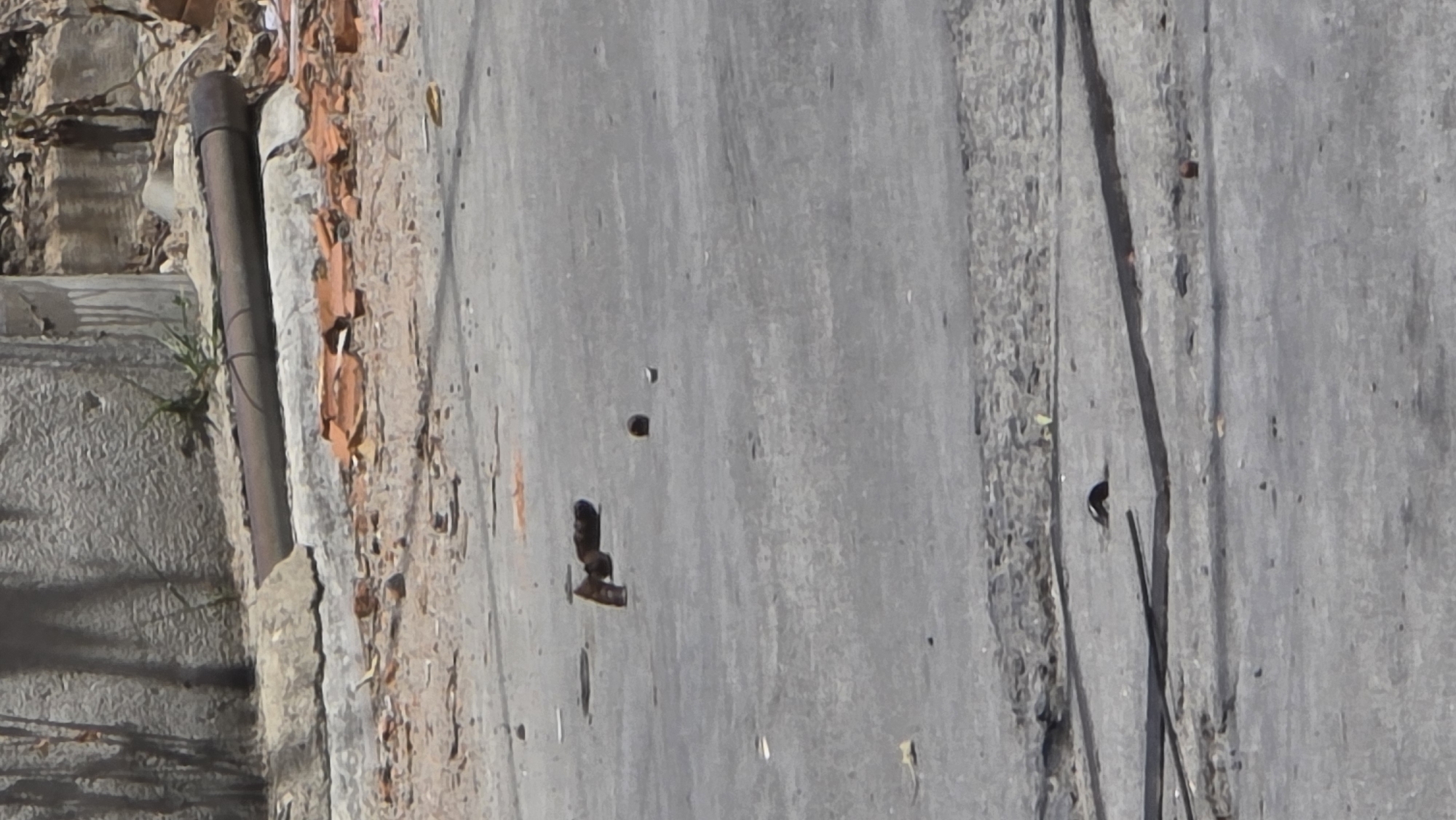 The image depicts a section of pavement in Bắc Nha Trang, Vietnam. The pavement is predominantly grey and shows signs of wear and damage, including cracks and small holes. Adjacent to the pavement, there is a patch of ground with broken tiles, a metallic pipe, and some vegetation. The scene appears to be outdoors in daylight.