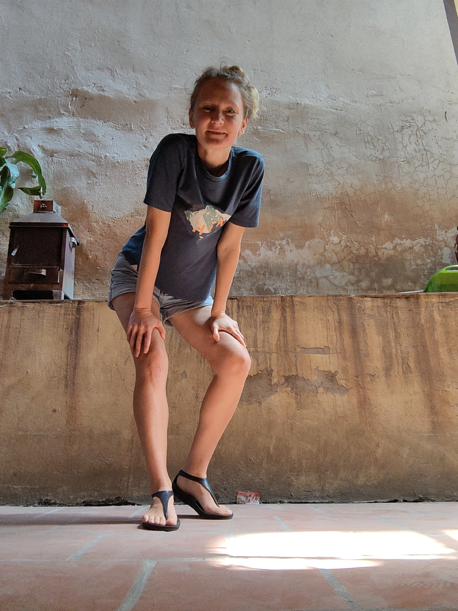 A young woman is depicted in a full-body shot, posing casually in what appears to be a covered outdoor area or hallway in Hà Nội, Vietnam. She is leaning forward with her knees slightly bent and her hands resting on her knees, smiling directly at the camera. She has light-colored hair pulled back and is wearing a dark t-shirt with a light-colored graphic on the front, light denim shorts, and dark T-strap sandals.
The background features a textured wall that shows signs of age, with visible cracks and areas of discoloration. The upper section of the wall is lighter, while the lower part is a warmer, earthy brown. A horizontal ledge runs across the mid-ground. On the left side of this ledge, a dark brown, ornate wooden object, resembling a lantern or small house, sits next to a green plant. A bright green object, possibly a bowl or pot, is partially visible on the right side of the ledge.
The floor consists of reddish-brown terracotta-style tiles. Strong natural light, likely sunlight, illuminates the scene, creating bright patches on the floor and distinct shadows, indicating a daytime setting. The woman's smile conveys a cheerful and relaxed emotion. There is no clearly legible text visible on her clothing or in the surroundings.
