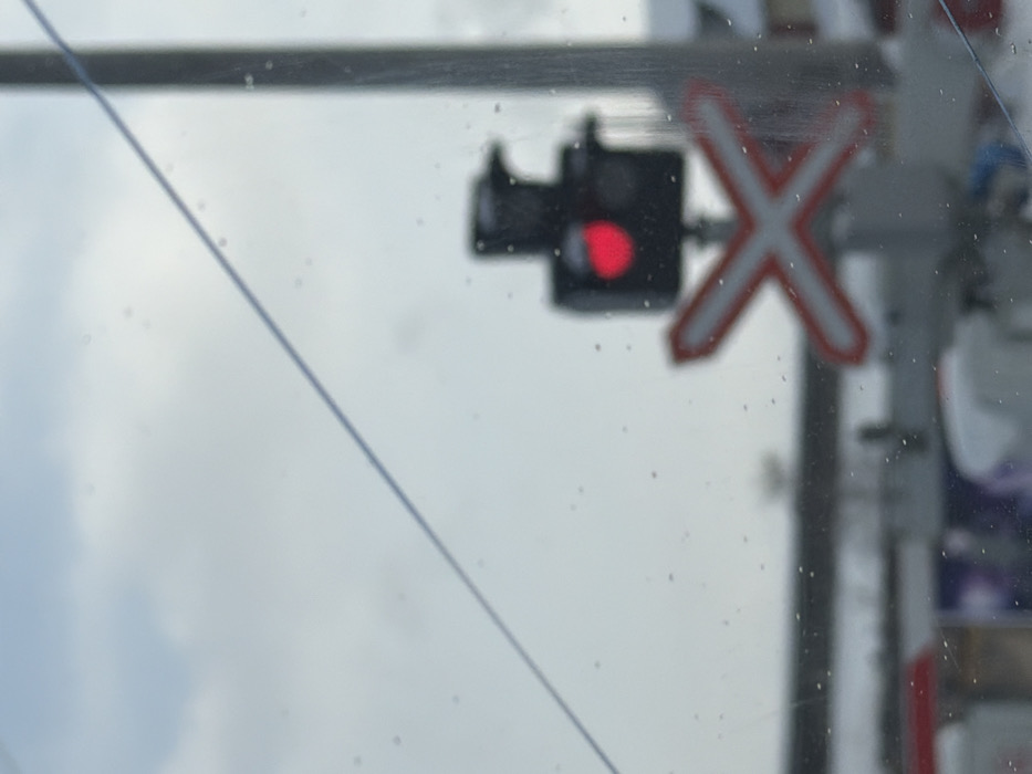 A blurred view through a glass surface reveals a railroad crossing signal. The signal features a red light illuminated in the middle of a black rectangular structure. To the right, a white and red "X" shaped crossing sign is visible, also out of focus. The background appears cloudy, possibly indicating overcast weather. A thin wire stretches diagonally across the frame. The glass has small water droplets dispersed on its surface.