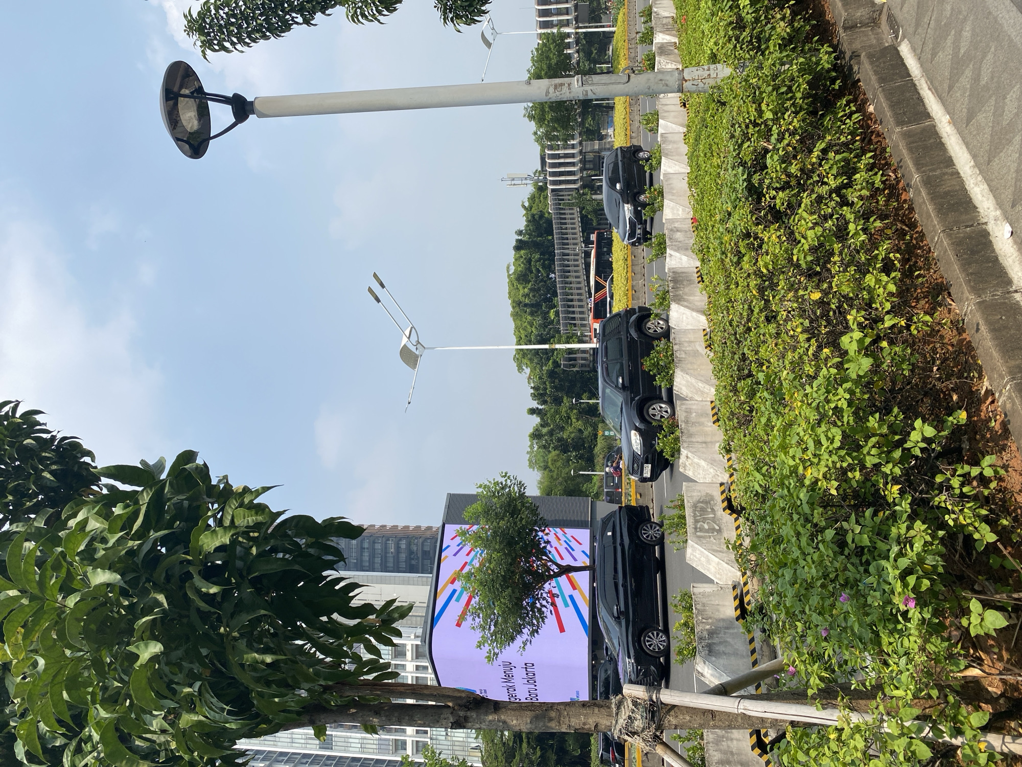 The image captures a street scene in South Jakarta, Indonesia, likely during daytime. In the foreground, lush green bushes and trees line a sidewalk. A tall, slender tree with dark bark stands on the left, partially obscuring a large LED billboard displaying colorful abstract shapes and text in Indonesian. The billboard is mounted on a modern building.
The street itself has multiple lanes of traffic. Several black SUVs are visible, driving in the same direction. A bus, possibly a public transport vehicle, is also in view, further down the road. A pedestrian bridge with a distinctive, curved architectural design spans across the road in the background, connecting buildings on either side.
Streetlights with a modern design are positioned along the road, with one prominent lamp post in the right foreground. The sky is clear with some wispy clouds, suggesting fair weather. The overall impression is of a busy urban environment with modern infrastructure and greenery integrated into the cityscape. The specific location is likely a main road or boulevard in a business or commercial district of South Jakarta.