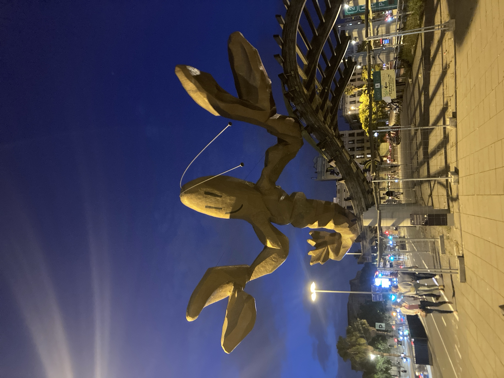 In Barcelona, Spain, at night, a large sculpture of a golden lobster with a smiley face is prominently displayed against a dark blue sky. The sculpture is supported by a dark, skeletal structure that arches overhead. In the foreground, a paved walkway is visible, with a metal fence bordering a street. Several people are walking on the sidewalk, and cars are driving on the street, their headlights and taillights illuminated. Traffic lights are visible in the distance, some showing red and others green. Trees and buildings can be seen in the background, with some of the buildings lit up. The overall scene suggests a public urban space at dusk or after dark, with the sculpture serving as a focal point.