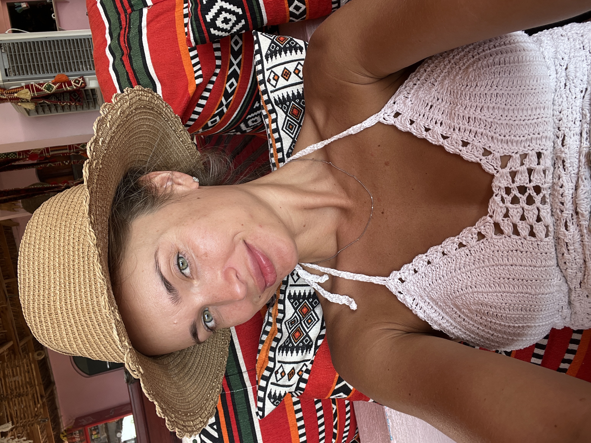 A fair-skinned woman with blonde hair and green eyes is taking a selfie. She is wearing a straw hat and a white crochet top. She is sitting on a couch with colorful striped pillows. In the background, there is a fan and a pink wall. The scene appears to be an outdoor cafe in the city of Nuweiba, Egypt, given the typical decor and climate suggested by the straw hat and outdoor setting.