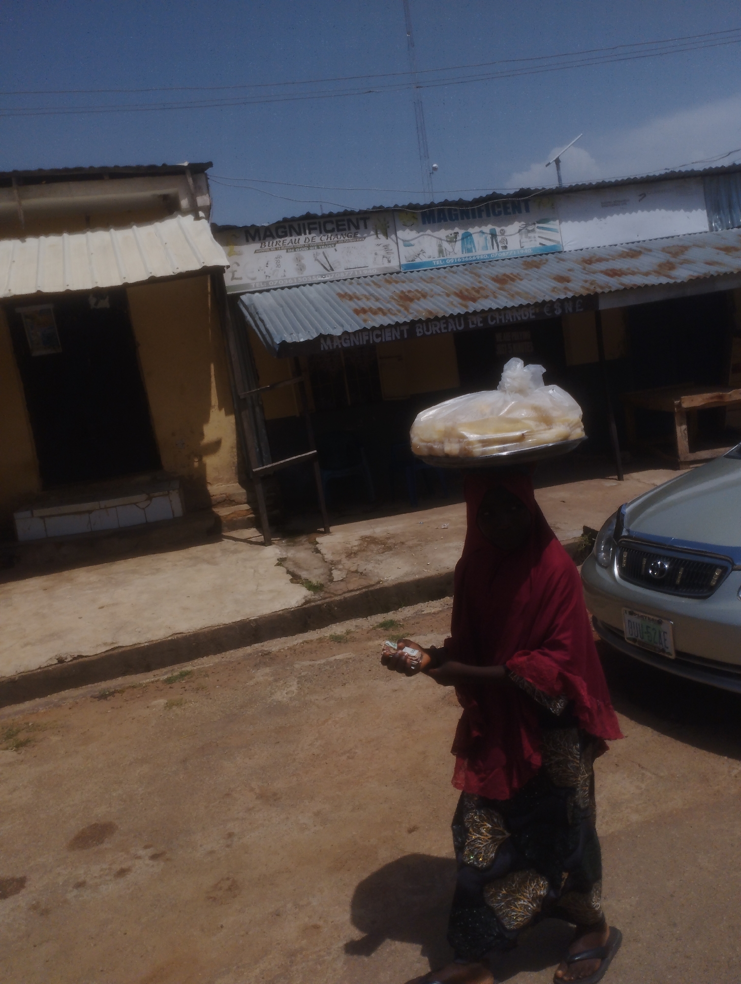 In a scene typical of a bustling Nigerian street, a woman, likely a vendor, carries a large platter of food items, possibly fried dough, balanced expertly on her head. She is dressed in a red headscarf and a patterned blue and gold skirt, with her hands outstretched, holding a wad of Nigerian Naira currency, suggesting she is engaged in a transaction or waiting for a customer. The setting appears to be an outdoor market or a commercial street in Jos, Nigeria.
Behind her, buildings with corrugated metal roofs and faded signage indicate a commercial area. One sign prominently displays "MAGNIFICENT BUREAU DE CHANGE," along with phone numbers, suggesting it is a currency exchange business. The architecture, with its simple structures and open-air feel, points to a warm, sunny day. The ground is a mix of paved concrete and dusty earth, with a visible drainage channel. A silver car with a Nigerian license plate (DUD-524E) is parked to the right, adding to the urban context. The overall impression is one of daily commerce and street life under a clear blue sky.