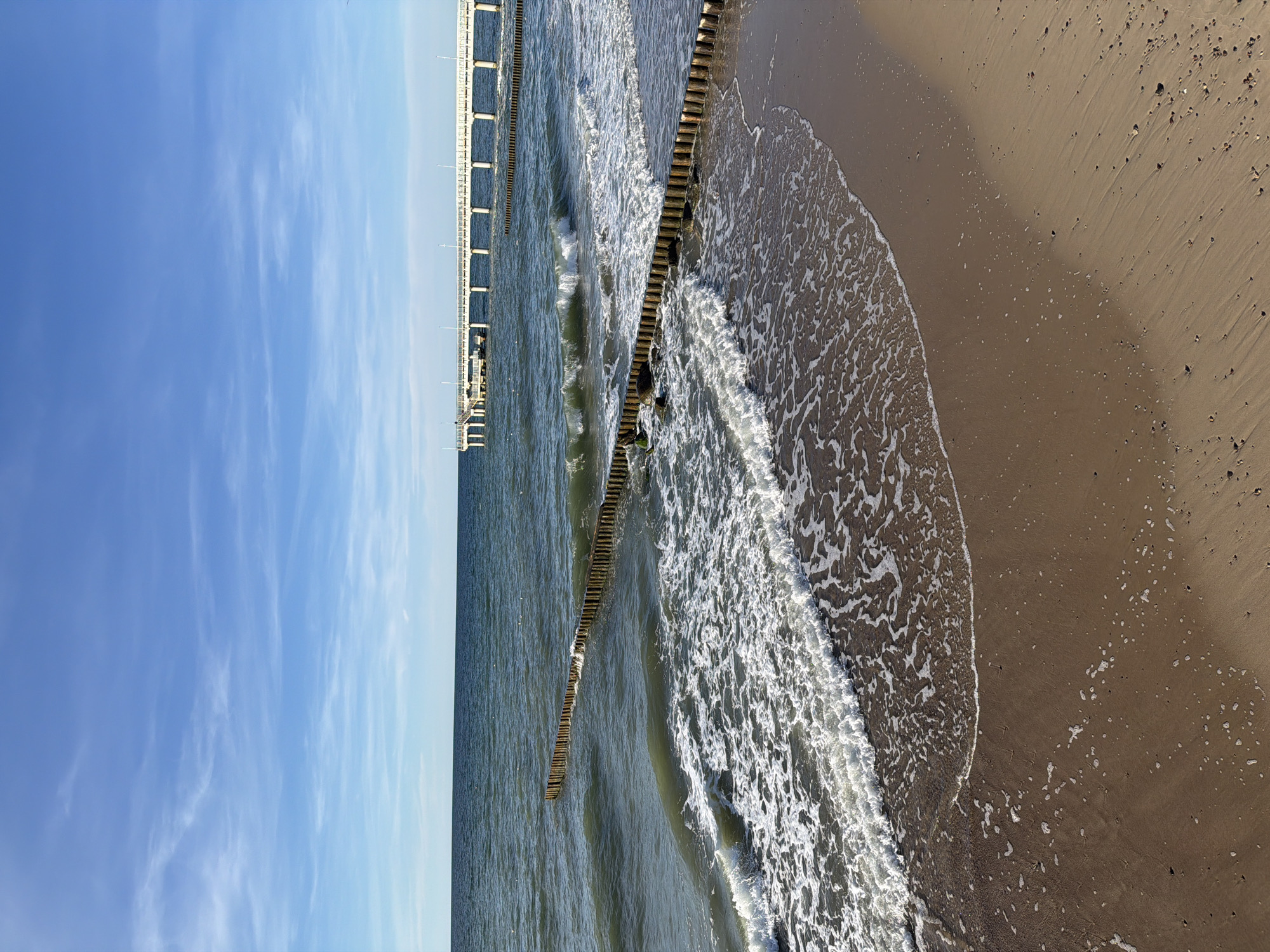 This is a photograph of the Baltic Sea coast in Zelenogradsk, Russia, featuring a beach, water, and a pier. The foreground is a sandy beach with some small shells and debris scattered across it. Waves are gently rolling onto the shore, leaving a foamy white surf line. Wooden groynes, appearing as lines of vertical posts, are partially submerged in the water, breaking the waves and creating calmer areas. In the background, a long white pier extends out over the sea, with a few scattered lights visible along its length. The sea is a deep blue-green, and the sky above is a clear blue with wispy white clouds. The overall scene is serene and depicts a typical coastal landscape.