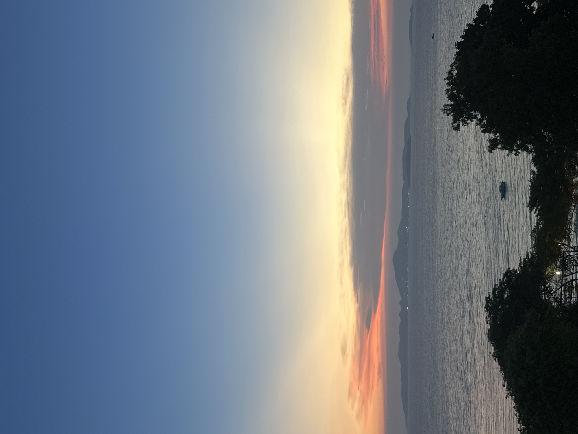 The image depicts a waterscape in Pattaya City, Thailand, at sunset or sunrise. The sky transitions from blue on the left to a hazy orange and yellow where the sun is setting or rising. The water fills the right half of the image, reflecting some of the sky's colors. There are hints of land in the distance, potentially islands or the mainland coastline. A small boat is visible on the water. Silhouetted trees frame the right side of the image.