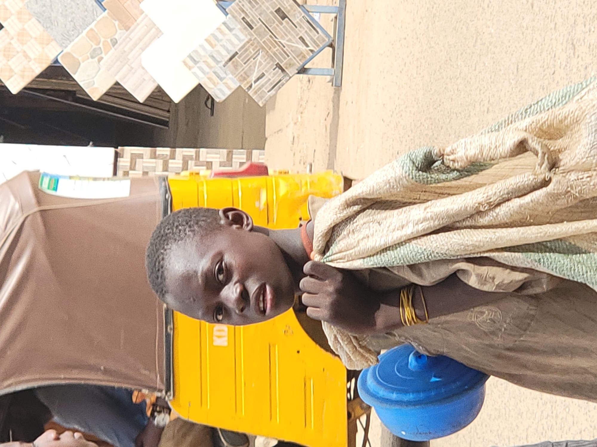 A young, dark-skinned person stands outdoors near what appears to be a storefront displaying tiles. The person is draped with a light-colored patterned cloth and has several gold-colored bracelets on their left wrist. A bright blue container rests near their left hip. A yellow structure, possibly a small vehicle, is visible behind the person. The setting is likely a commercial area in Maiburiji, Nigeria, based on the prompt.