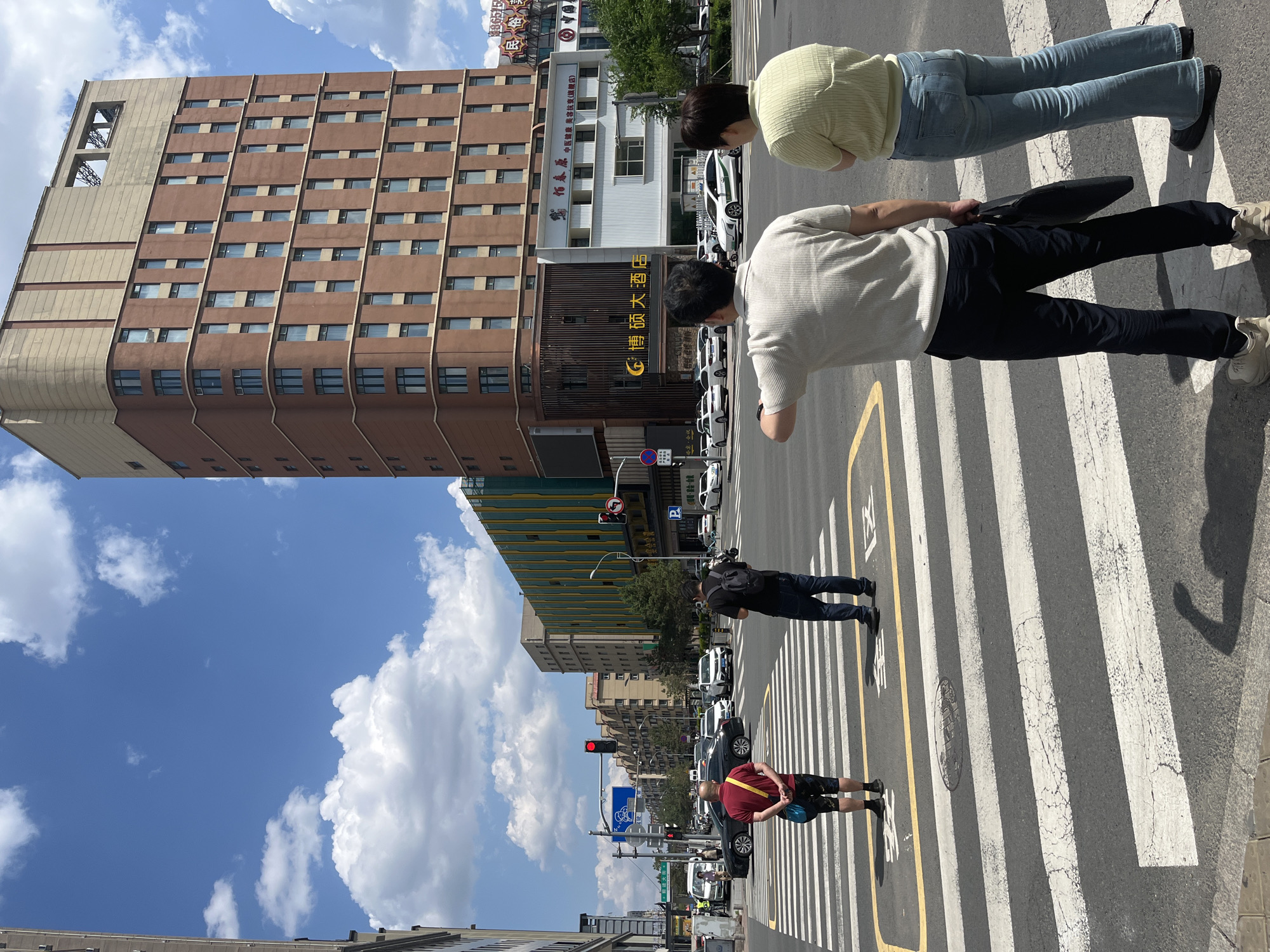 The image captures a scene in Changchun City, China. Pedestrians are crossing the street on a crosswalk. There are four discernible individuals: two in the foreground and two further down the crosswalk. The building is identified as 'Bo Shuo Grand Hotel'. Other buildings are visible in the background. The sky is blue with scattered white clouds. Multiple vehicles are on the road and the traffic lights indicate red, suggesting that vehicles are stopped.