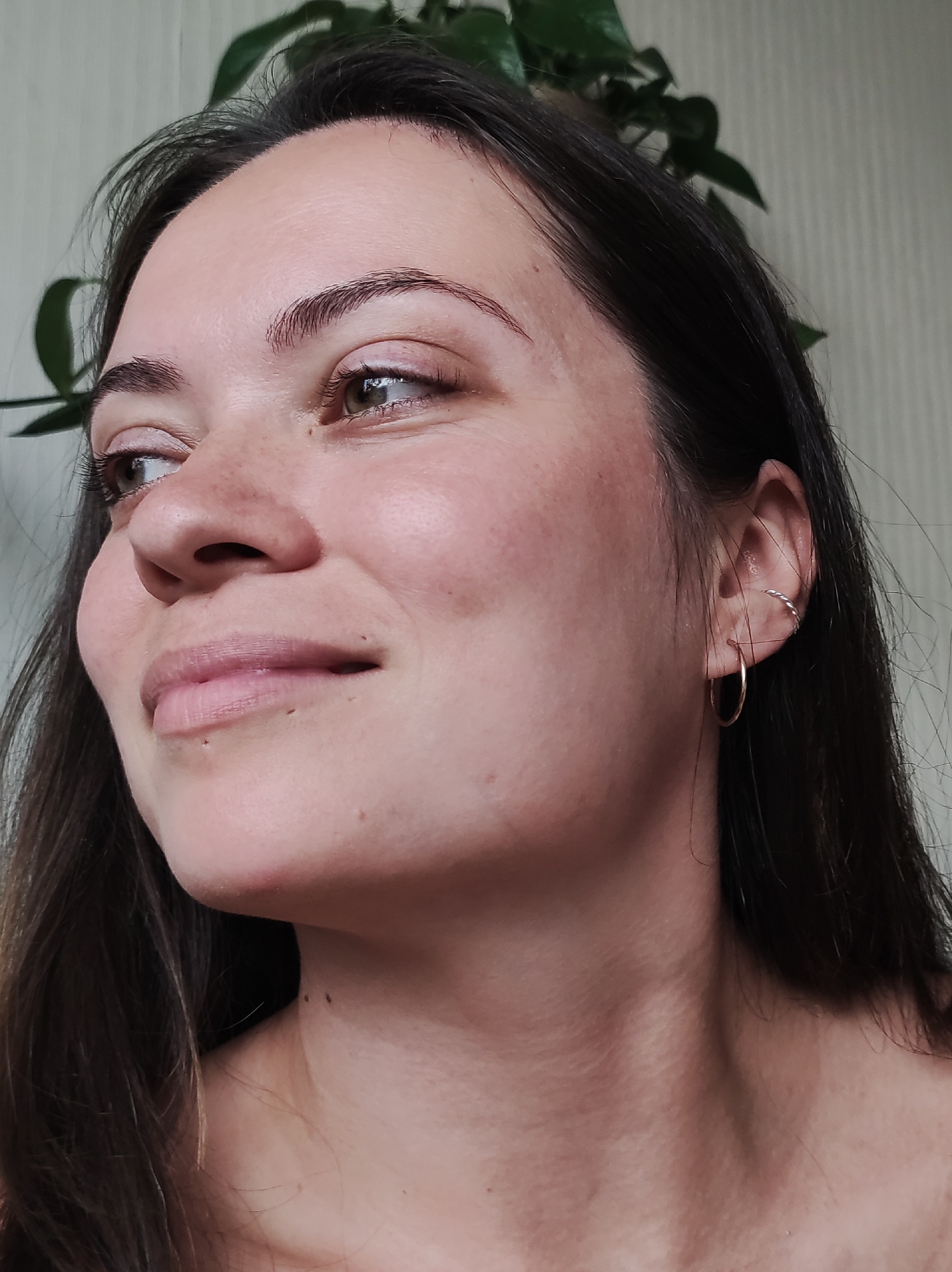 A close-up, profile shot shows a young woman with dark hair and brown eyes. She has fair skin with freckles and appears to be wearing subtle makeup. She is looking off to her left, with a gentle smile. She has a hoop earring in her left ear. In the background, out of focus, are green leaves of a plant and a textured wall. The image is a digital photograph. There is no information to determine its location in Chaikovsky, Russia.