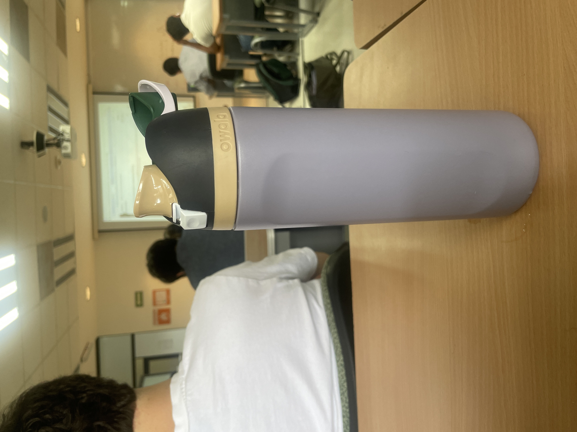 A light purple Owala brand water bottle stands upright on a light brown wooden desk, serving as the central focus of the image. The bottle features a black cap with a beige band and distinct green and white elements around its sip and straw mechanisms.
The scene depicts an indoor classroom or lecture hall environment, with the perspective seemingly taken from behind an individual seated at a desk. The top of this person's head and the back of their white shirt are visible in the foreground. In the background, several other individuals, likely students or attendees, are seated at similar desks, mostly viewed from behind. They wear light-colored attire, and some have dark hair.
A projector screen, displaying some content that is not clearly legible, is visible on a far wall. The room is illuminated by overhead fluorescent lights and features light-colored walls. Bags are discreetly visible tucked under some of the desks in the background. The overall impression is that of a daytime academic setting, possibly during a lecture or class in San Andrés Cholula, Mexico. No specific emotions or weather conditions are discernible from the image.