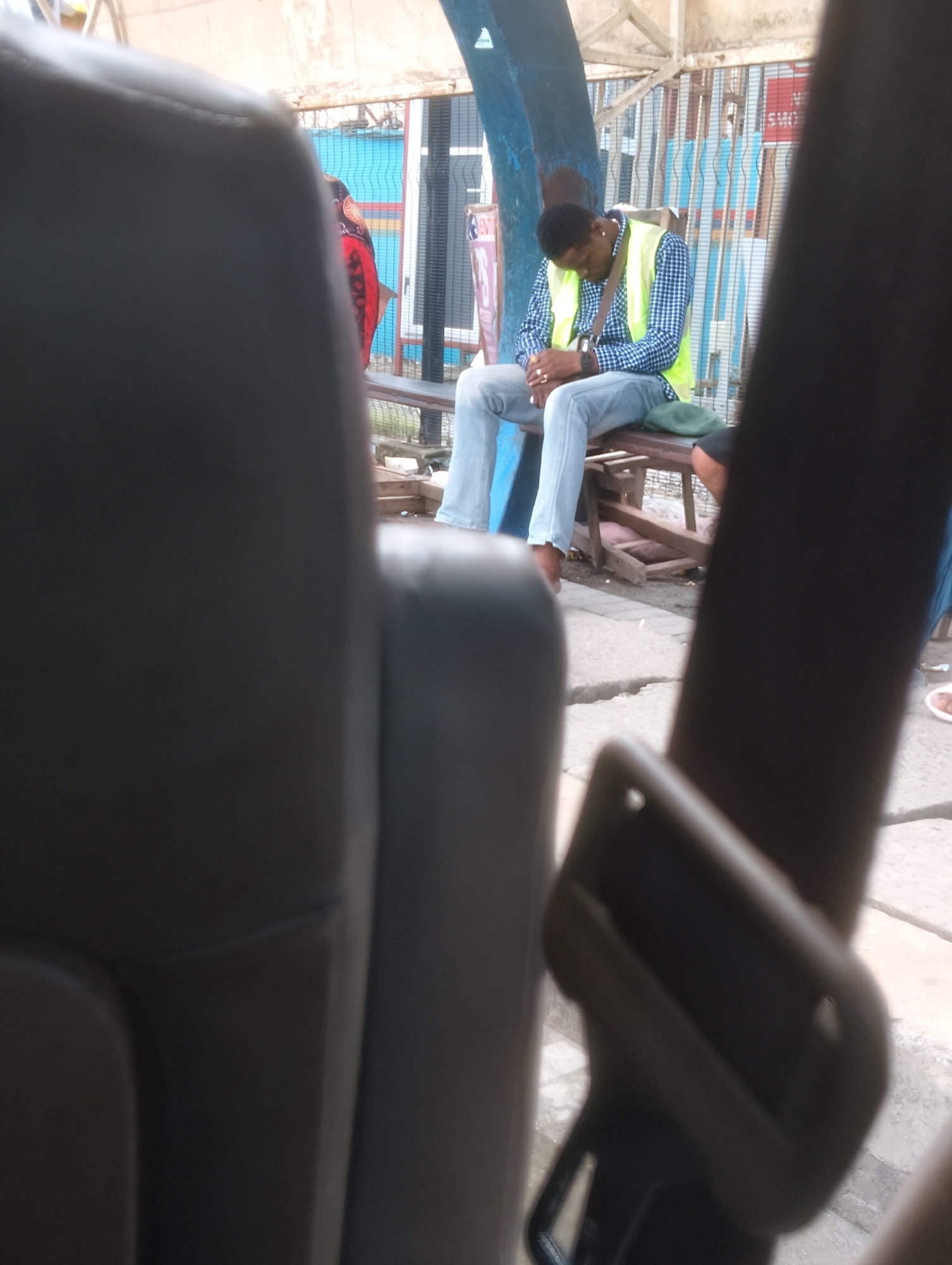 In Oworonshoki, Nigeria, a Black man wearing a blue and white checkered shirt and a high-visibility yellow vest sits on a wooden bench, his head bowed as if sleeping. He is wearing blue jeans, a watch, and rings on his fingers. A green cap rests on the bench next to him. Behind him, a blue column supports an overhead structure. The background features a corrugated metal fence with vertical blue and red stripes. The foreground is dominated by the blurry interior of a vehicle, showing dark gray seats and a black seatbelt mechanism.