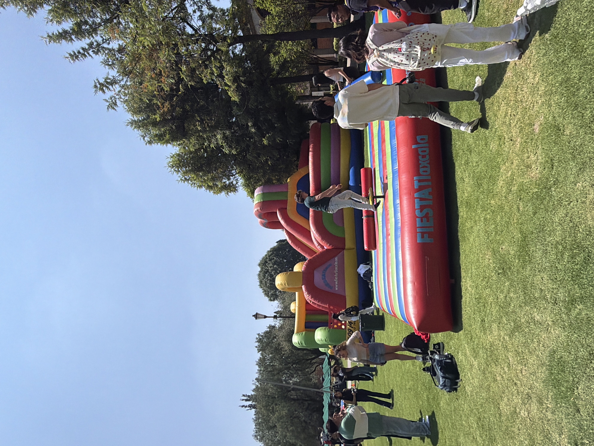 The image depicts an outdoor event on a grassy area, possibly a park, under a clear, blue sky. A large, colorful inflatable obstacle course, labeled "Fiesta Tlaxcala," is prominent in the scene. People of various ages are interacting with the inflatable, with some actively jumping on it and others standing around observing. Trees surround the area, providing a backdrop. Based on the text on the inflatable, the location is possibly Tlaxcala in Mexico.