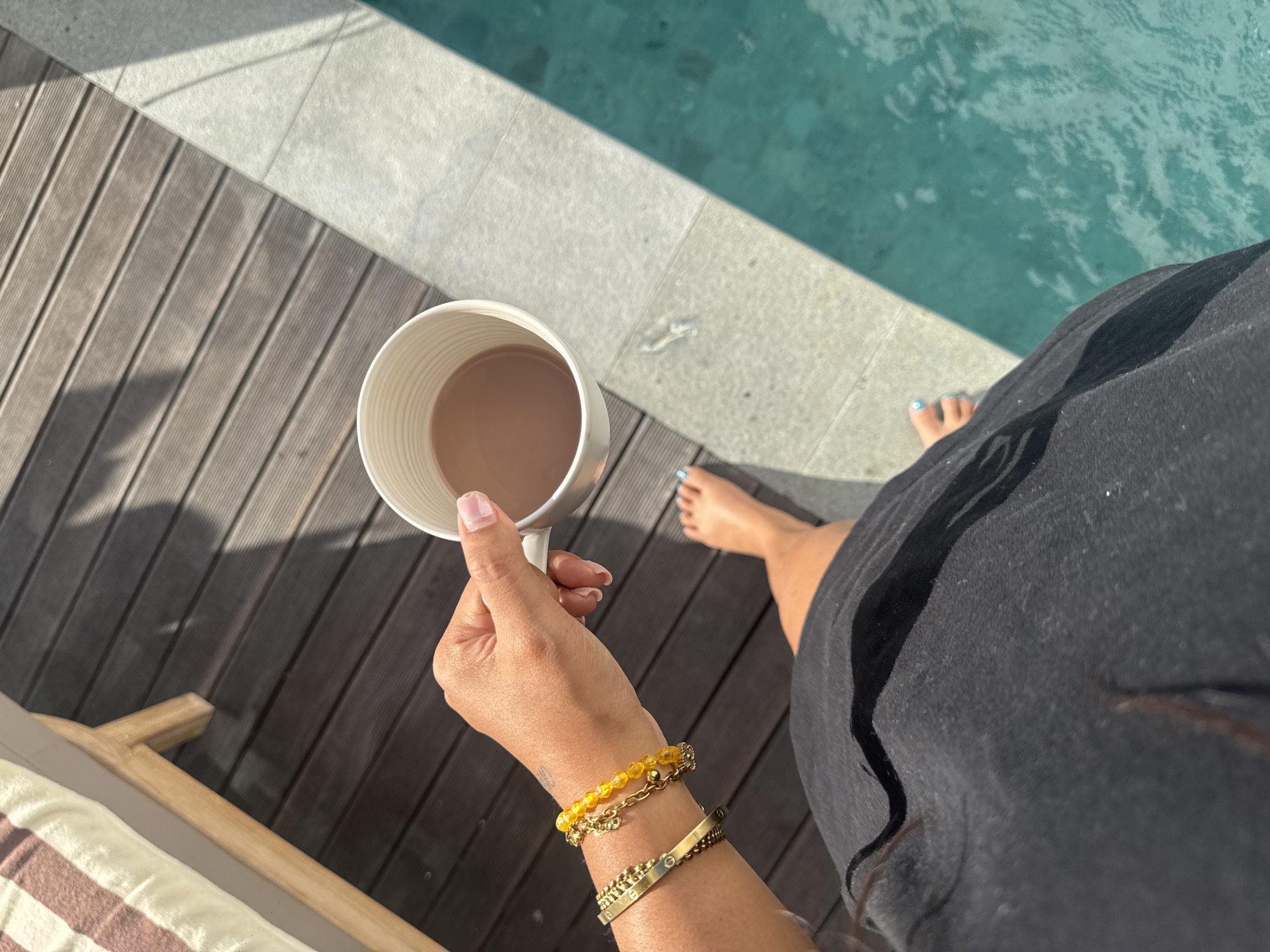 A top-down perspective shot captures a person holding a white mug of coffee by a swimming pool. The scene is set on a wooden deck next to a turquoise pool. The sun is casting distinct shadows, indicating it is a sunny day, likely mid-morning or afternoon. The person is wearing a black garment and adorned with several bracelets on their wrist. Their feet, with blue nail polish, are visible near the pool's edge, suggesting relaxation and leisure. The presence of a lounge chair with a striped cushion in the foreground hints at a vacation or resort setting. The overall mood is tranquil and relaxed, conveying a sense of enjoyment and peacefulness. The location is identified as North Kuta, Indonesia.