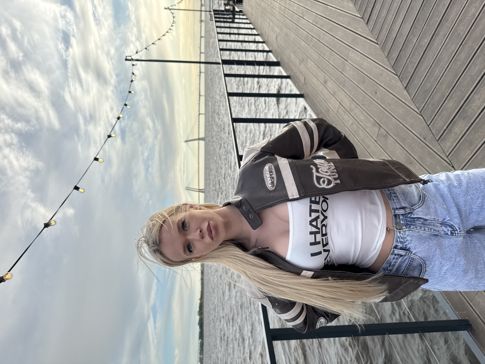 A woman with long blonde hair stands on a wooden pier, wearing a white tank top with "I HATE EVERYONE" printed on it, a brown and white racing jacket, and light-wash denim jeans. She has a navel piercing. In the background, a wide body of water stretches to the horizon, with a large bridge visible in the distance. String lights are hung above the pier. The scene appears to be located on Krestovsky Island in Saint Petersburg, Russia, possibly near the Western High-Speed Diameter (WHSD).