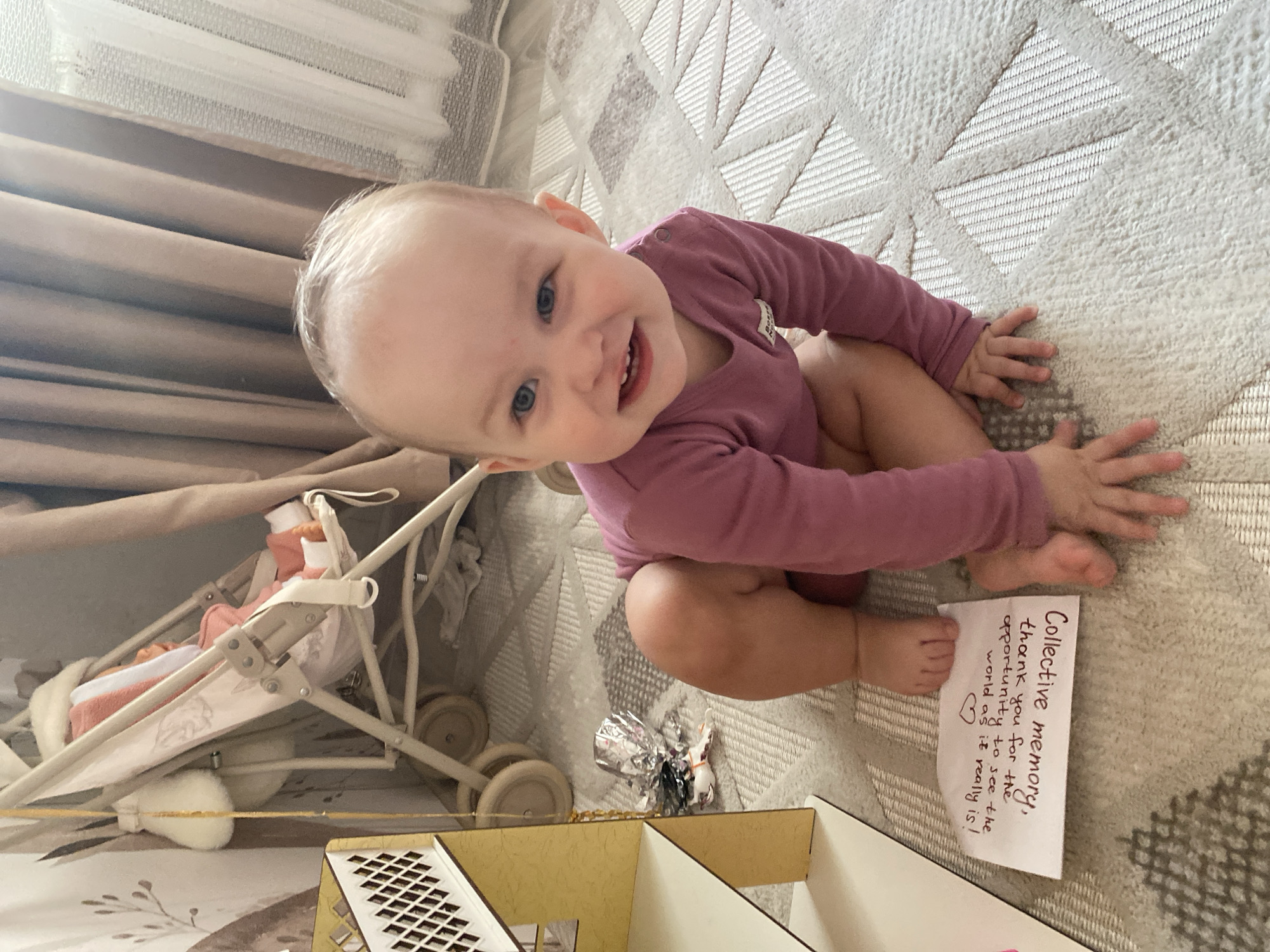 An indoor scene in Chaikovsky, Russia, features a smiling young child, appearing to be a toddler, positioned centrally on a light-colored rug with a geometric pattern. The child has light hair, blue eyes, and is wearing a long-sleeved magenta bodysuit, with bare legs and feet, crouching forward with hands flat on the rug. The child's mouth is open in a wide smile, directed towards the viewer. To the child's immediate left, a white piece of paper with handwritten text reads: "Collective memory, thank you for the opportunity to see the world as it really is! <3". Further to the left, a light-colored wooden or cardboard dollhouse or shelving unit is visible, featuring a white lattice design on its upper left side. Behind this unit, a beige-colored toy stroller contains a doll wrapped in an orange-pink blanket. Crumpled silver foil is present on the rug near the base of the dollhouse. The background shows brown opaque curtains and white sheer curtains, indicative of a window. The setting is a domestic interior.