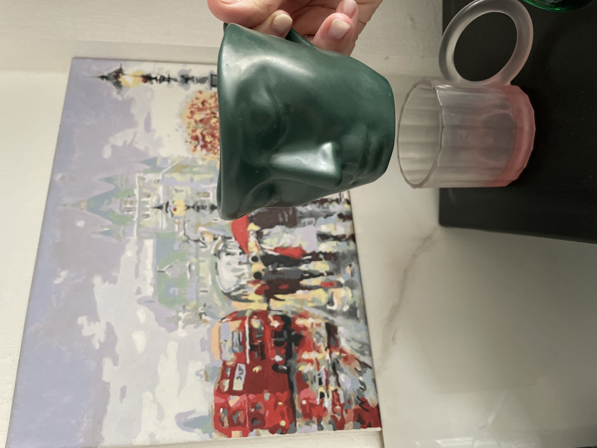 A painting depicting a European cityscape is in the background. Dominating the lower portion of the painting are red double-decker buses. Above this are pedestrian figures in shades of red, white, and grey. In the top portion of the painting is a grey building or monument. In the foreground, a hand holds a green mug. To the right of the green mug sits a transparent mug with a pink tint, resting on a black surface. The environment consists of a plain, white wall. No specific information indicates the location is Krasnoyarsk, Russia.