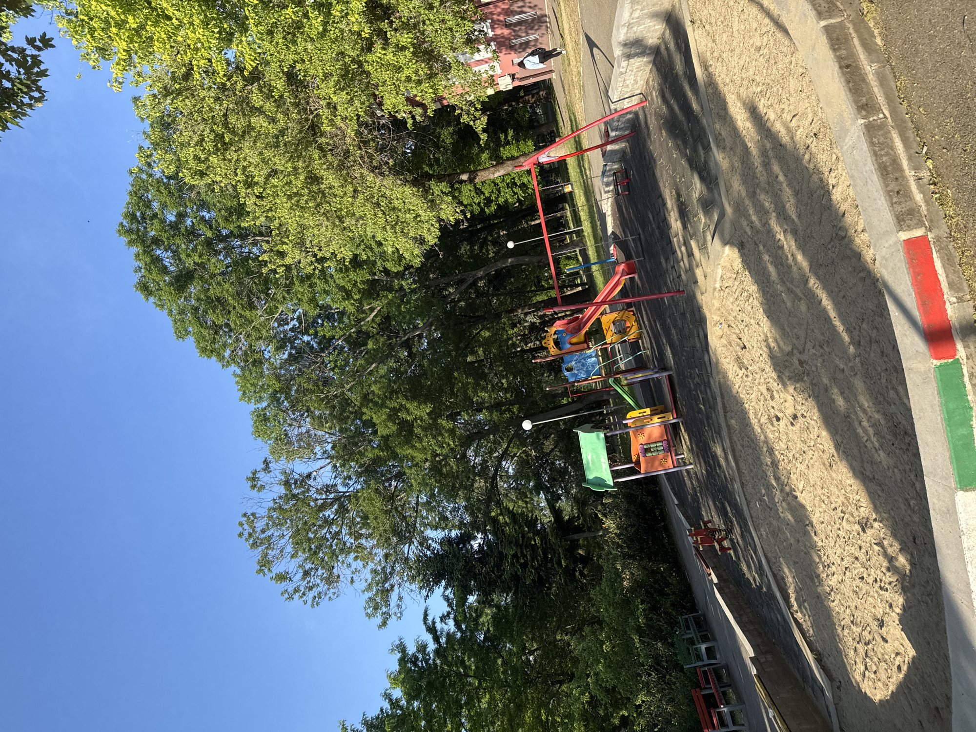 The image depicts a playground in Buhovo, Bulgaria. It features a sand-filled play area surrounded by concrete edging marked with red and green paint. Playground equipment including a slide and various climbing structures are visible. Trees provide shade over the playground. A building is visible in the background.