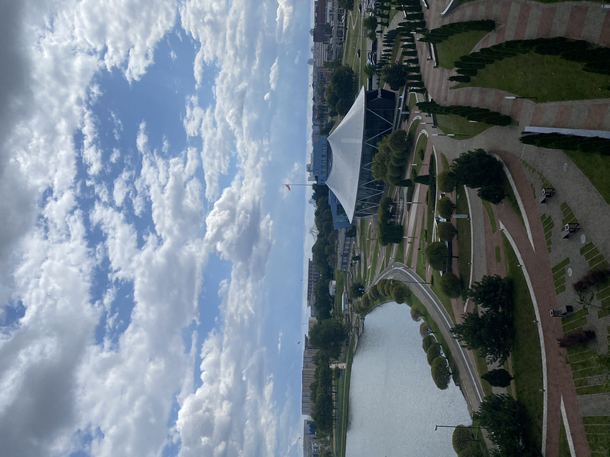 An aerial, wide shot captures a panoramic view of a park area in Minsk, Belarus, adjacent to a body of water. The sky is a mix of blue and scattered white clouds. In the foreground, paved pathways with embedded lighting and green grass areas with manicured trees form a park landscape. A curved road runs alongside the water. Further back, a modern building with a white, tent-like roof stands out, a flag flying from a pole in front of it. Beyond this, several larger, rectangular buildings are visible, some with flags, and a cityscape with more structures stretches into the distance. The overall scene suggests a well-developed urban park with recreational and architectural elements.