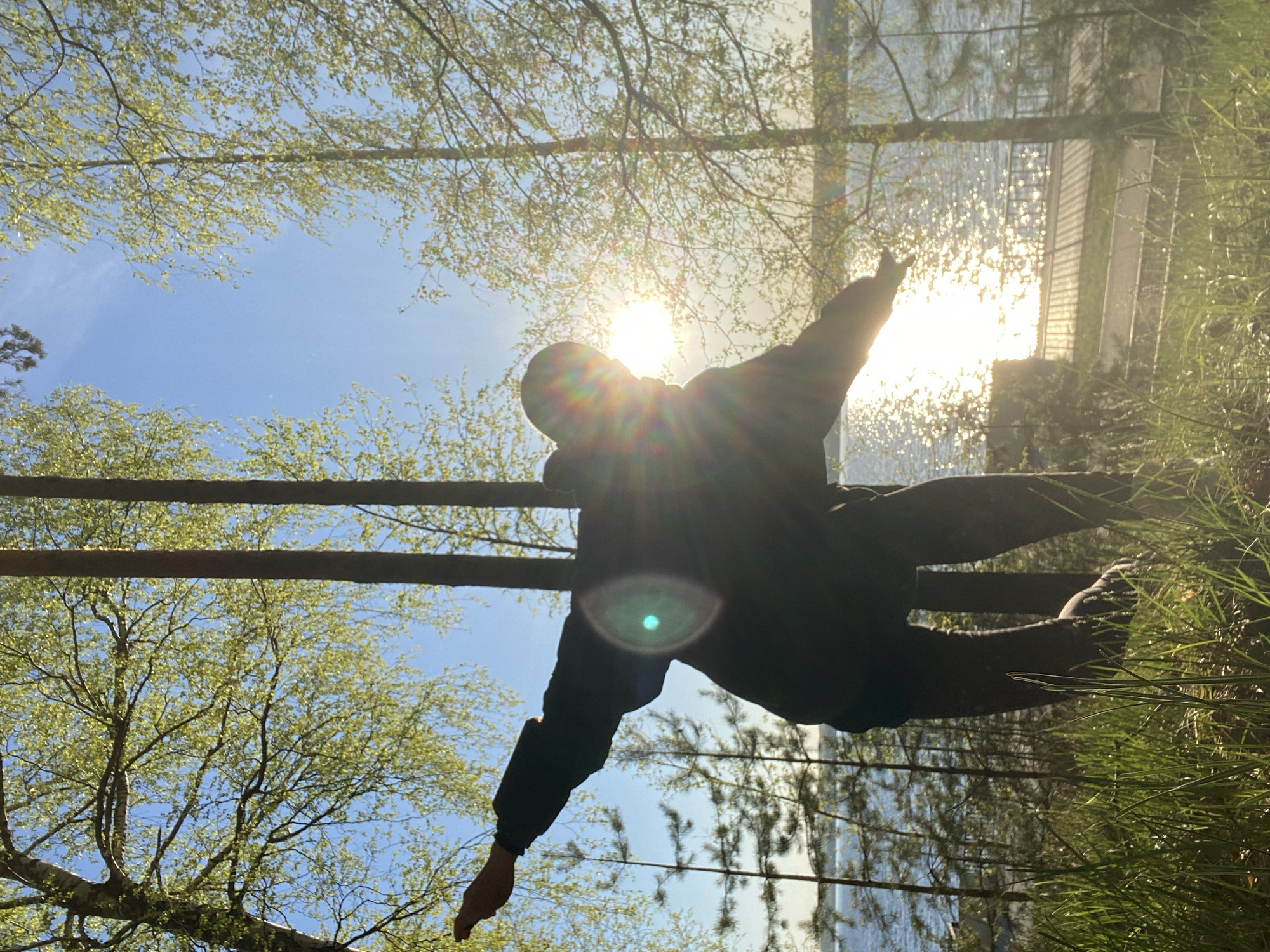 A person is captured mid-action, appearing to be jumping or balancing between two parallel wooden beams. The scene is situated outdoors with lush green grass on the right and a body of water, likely a lake or river, reflecting sunlight and the silhouette of a building in the background. The sky is a clear blue with dappled sunlight filtering through the tree branches above. The person is wearing dark clothing, making it difficult to discern details. The location is Chaikovsky, Russia.