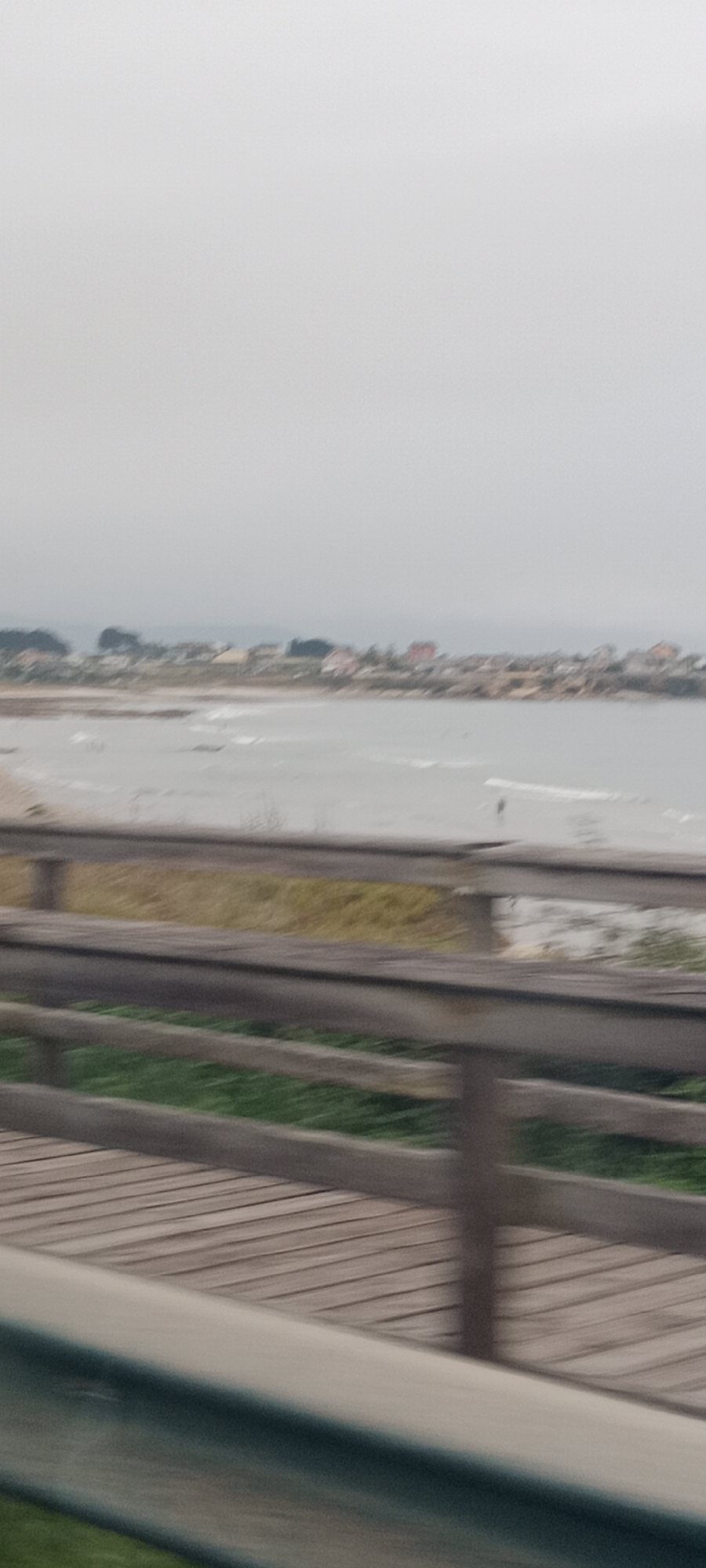 The image shows a blurred view of a wooden boardwalk or pier in the foreground, with a grassy area and a wooden railing. Beyond the boardwalk is a beach and the sea, with gentle waves. In the distance, a line of buildings, possibly houses, is visible along the coastline, suggesting a small town or village. The sky is overcast and hazy. The scene appears to be the coastal area of Reinante-praia, Spain, on a cloudy day. There are no discernible people or specific actions occurring.