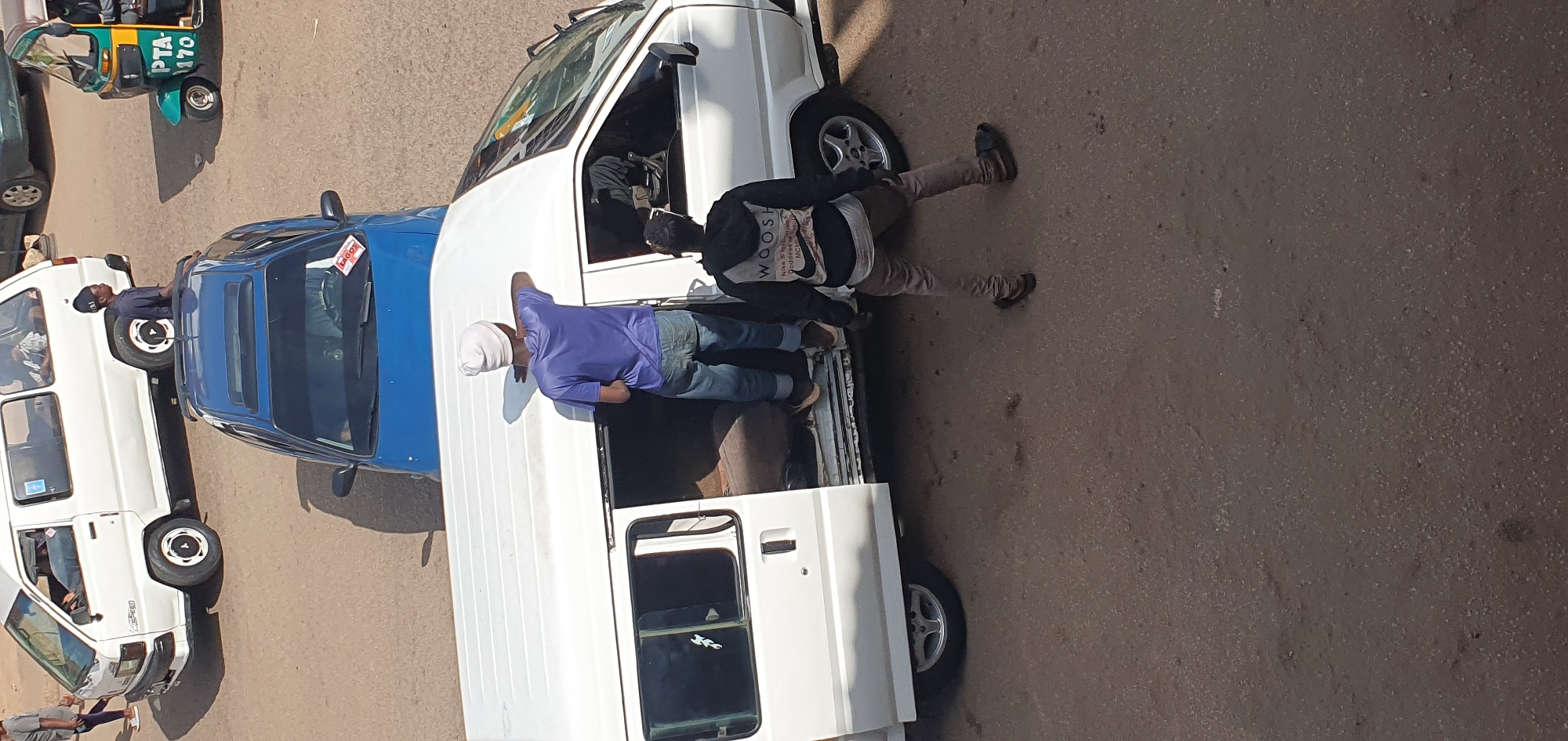 In Bukuru, Nigeria, a white mini-van is parked on a paved road. Two individuals are near the open side door of the van. One person, wearing a white head covering, a purple t-shirt, and blue jeans, is standing on the van's roof, with their right hand on the roof and their left arm reaching towards the roof. Another person, wearing a black hooded jacket with "WOOSH" printed on the back, grey pants, and dark sandals, stands facing the van. The jacket has a Nike logo and text below it. The road surface is visible, with asphalt and some dirt. In the background, a blue car with a sunroof is parked behind the white van, and further back, a green and yellow auto-rickshaw is visible. Another white van is parked to the left of the blue car. A person wearing a dark blue shirt and a black beanie is sitting on the roof of the white van on the left.