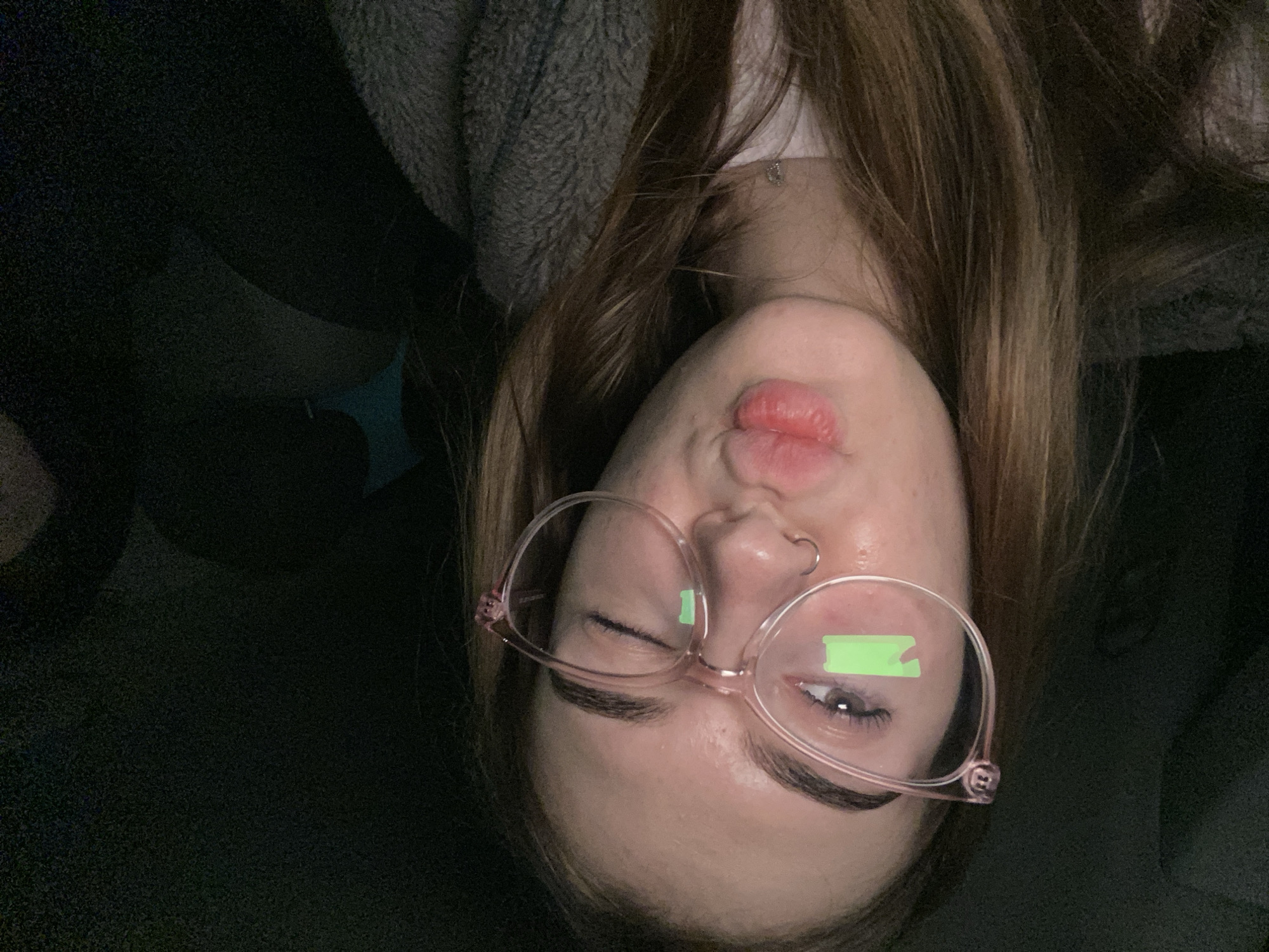 A close-up, tilted selfie captures a young person, likely a woman, in a dimly lit indoor setting. The subject has long, brown hair and is wearing a grey, textured garment, possibly a hoodie. They wear light pink, translucent-framed glasses. On each lens, a distinct, rectangular, light green glowing shape is visible, appearing as a reflection from a screen or a digitally added element. One eye is partially closed or squinting, while the other is visible through the lens. The lips are pursed. A small silver hoop nose piercing is visible on the nostril, and a faint silver chain or necklace can be seen around the neck. The background is dark and indistinct, with a subtle hint of a bluish-teal object in the lower left, reinforcing the low-light ambiance. No visible text is present in the image.