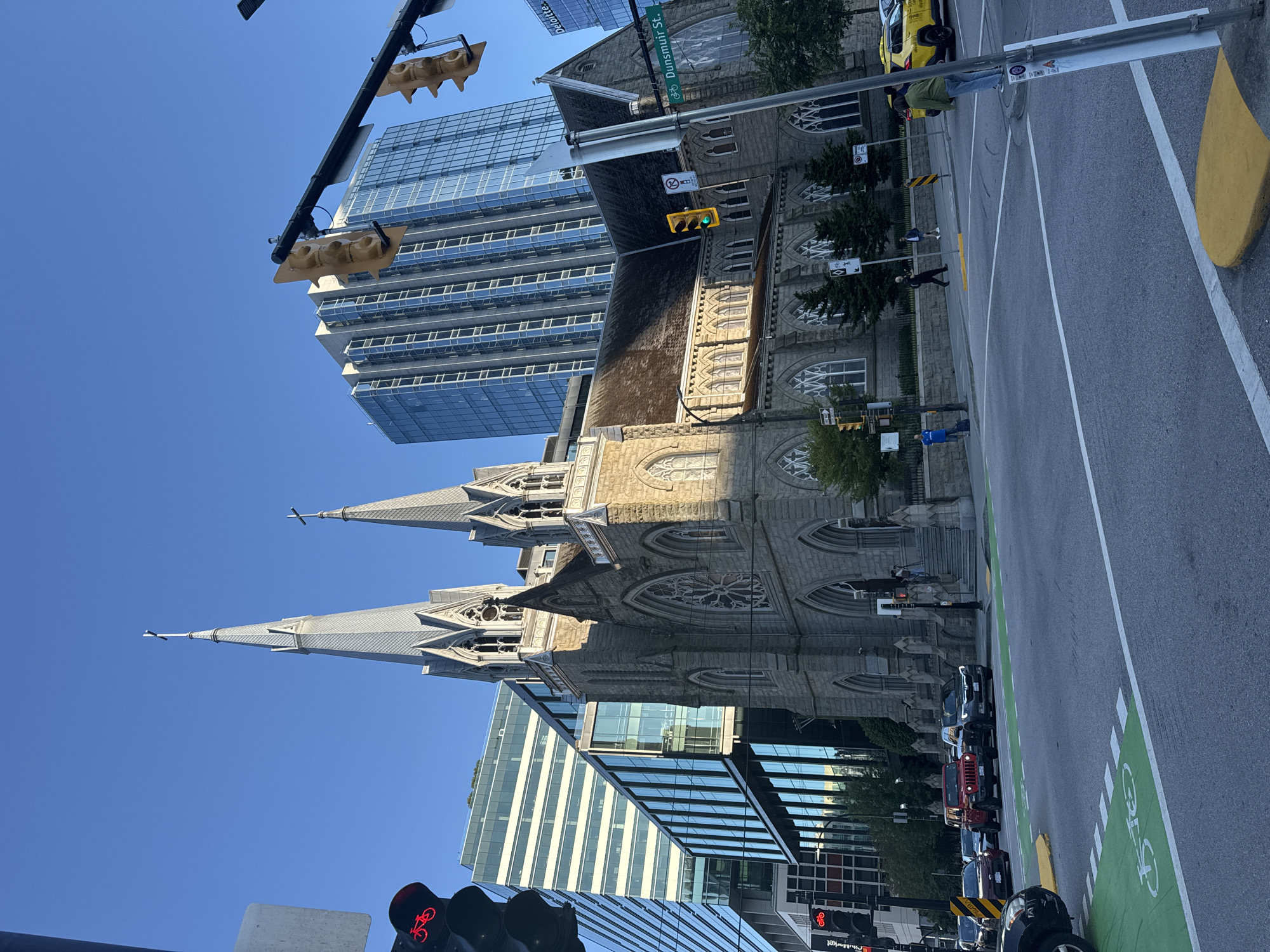 The image depicts an urban street scene in Vancouver, Canada, featuring a historic stone church with two prominent spires. The church is situated next to modern glass-fronted buildings. Traffic lights, street signs, and marked bike lanes are visible on the asphalt road. Several vehicles, including cars and a yellow taxi, are present. Pedestrians can be seen walking on the sidewalk. The sky is clear and blue, suggesting a daytime scene. The street sign indicates "Dunsmuir St."