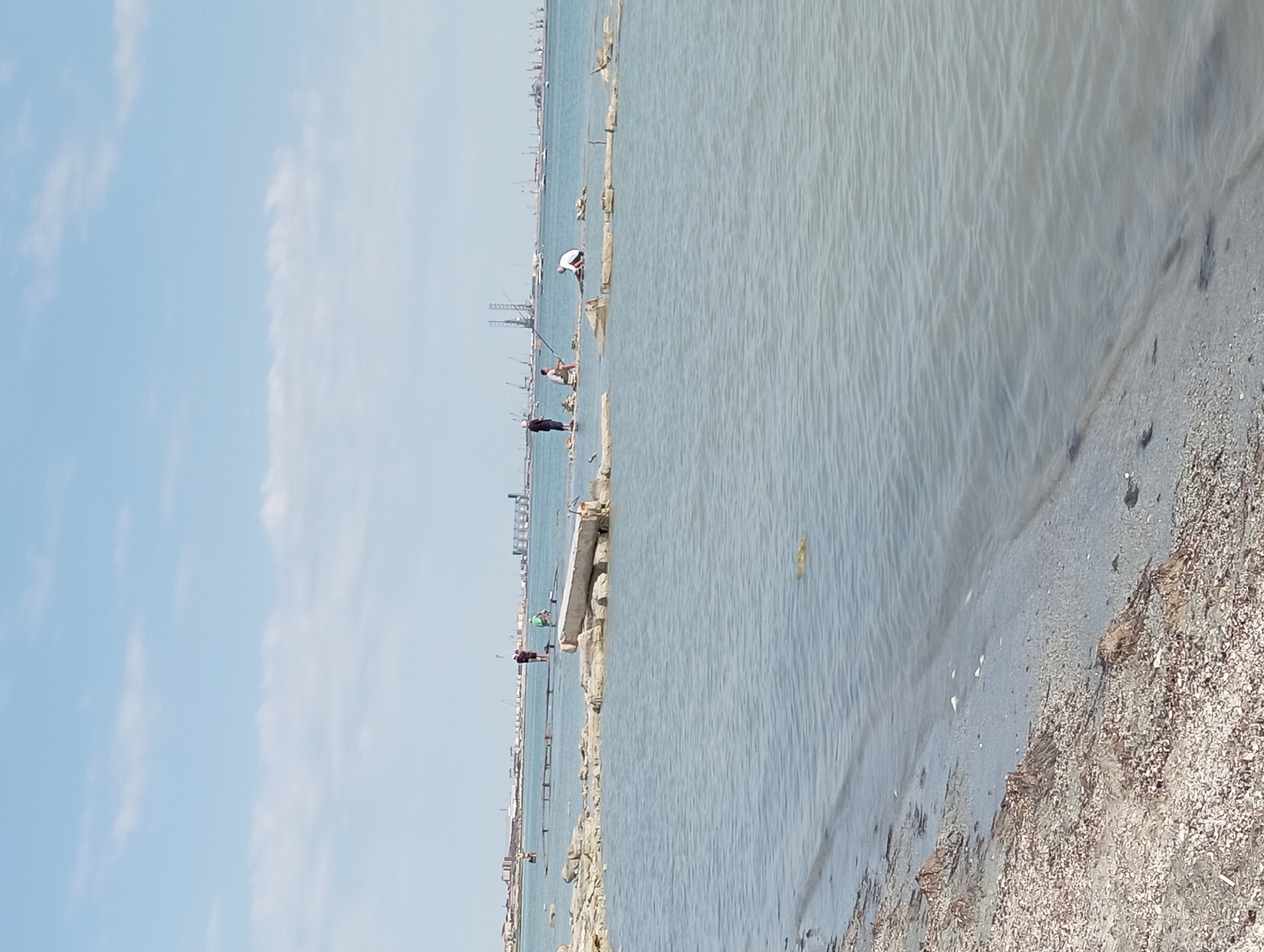 The image shows a waterfront scene in Aktau, Kazakhstan, with the Caspian Sea in the foreground. The beach is covered with shells and debris. Several people are visible in the shallow water, some fishing and others wading. In the background, there is a distant industrial area with cranes and port facilities, suggesting a coastal city with active maritime trade. The sky is partly cloudy.