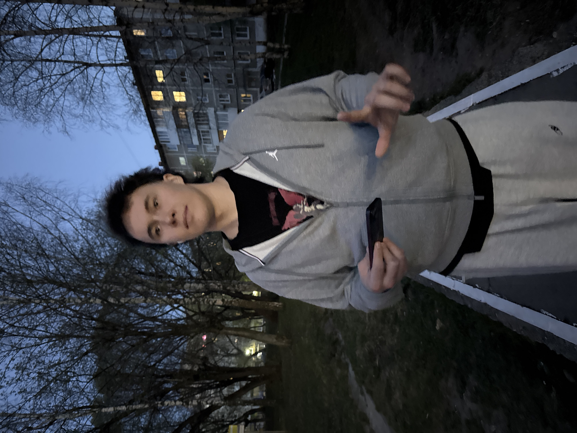 A young man stands outside in Perm, Russia. He wears a gray Jordan brand zip-up hoodie over a black t-shirt, and gray sweatpants with a black waistband. He holds a black cell phone in one hand and gestures with the other. Bare trees and a multi-story building with lit windows are visible in the background under a blue evening sky. The location appears to be on a grassy area next to a sidewalk.