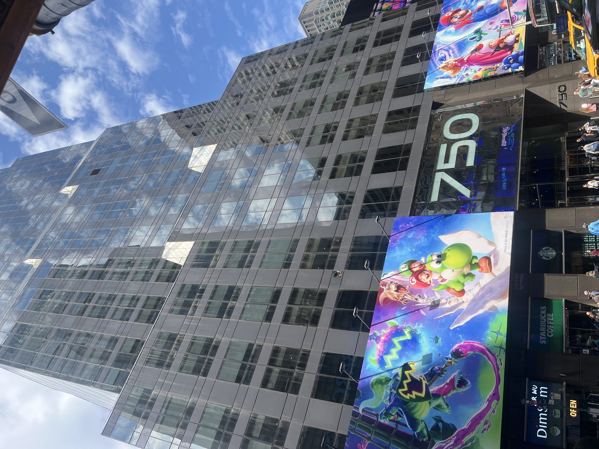 Here is a description of the image:
The image captures a bustling street scene in New York City, focused on a towering glass building adorned with large electronic billboards. A prominent billboard displays imagery from a "Super Mario" film, with a yellow taxi nearby. Additional billboards for Starbucks Coffee and Dim Sum WOO are also visible. The building reflects the cloudy blue sky, adding to the visual depth. The number "750" is prominently displayed on the building's exterior, near the billboards. People are walking by on the sidewalk below, though the focus is on the architectural details and advertisements.