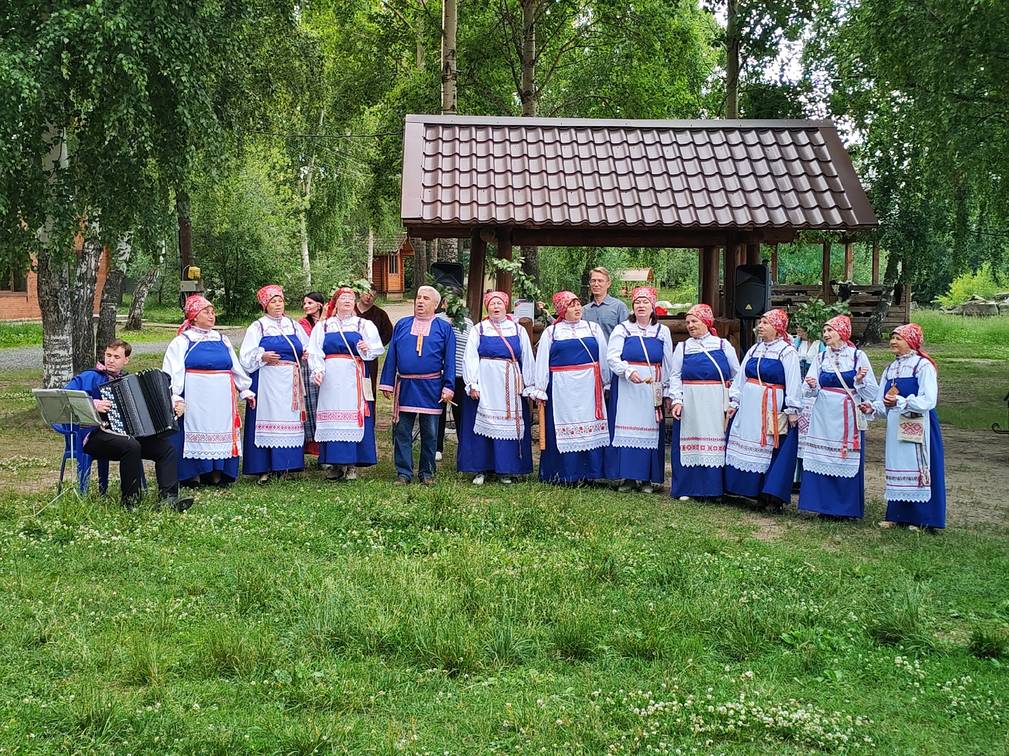 A group of approximately 14 people, dressed in traditional Russian folk costumes, are gathered outdoors in a park-like setting. Most of the group are women wearing blue dresses with white embroidered aprons and red headscarves. There are also at least two men, one in a blue tunic with embroidered trim and dark pants, and another man in a blue shirt standing behind the women. To the far left, a man is seated on a blue chair playing an accordion. Behind the group is a wooden structure with a brown tiled roof, resembling a gazebo or small pavilion. The ground is covered in green grass with scattered white flowers. The background consists of lush green trees, suggesting a natural or recreational area. The overall scene depicts a cultural performance or gathering. The image does not contain specific indicators to precisely pinpoint the location within the city of Chaikovsky, Russia, other than the general suggestion of a park.