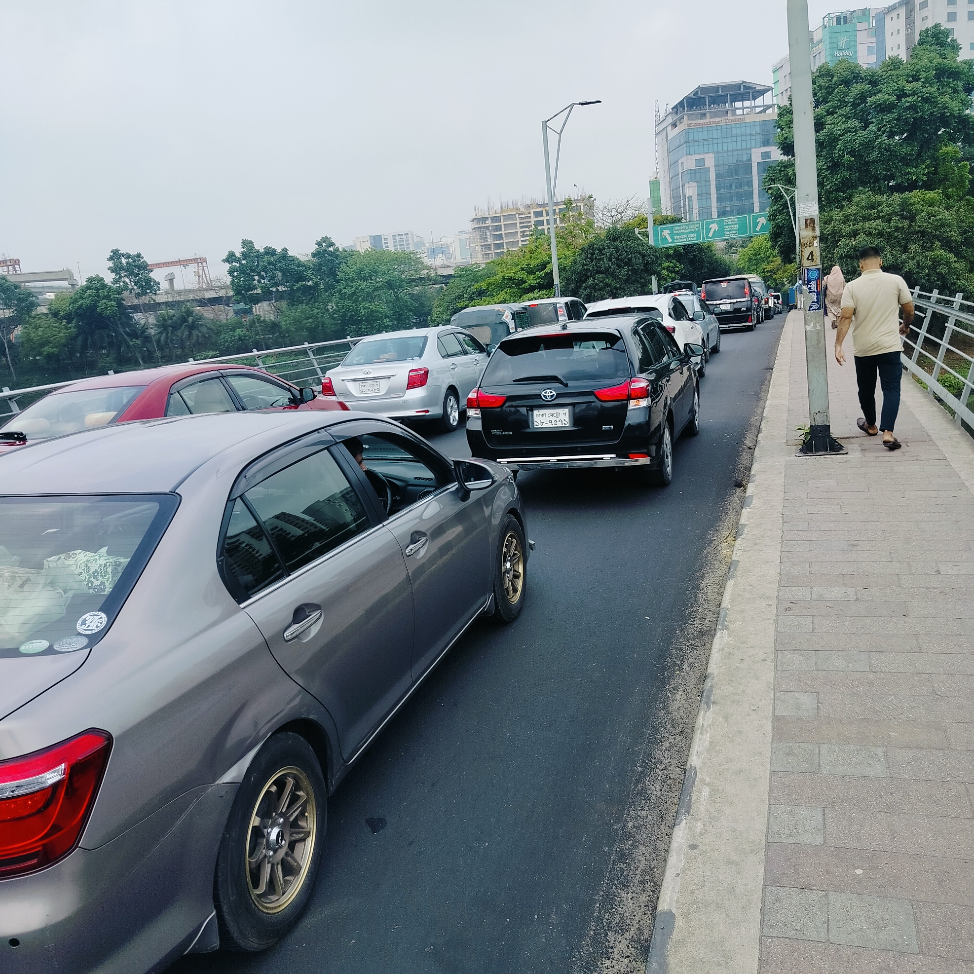 The image shows a congested street in Dhaka, Bangladesh. Several cars are stuck in traffic, including a grey Toyota Corolla Axio in the foreground and a black Toyota Corolla Fielder behind it. A red sedan and a silver sedan are also visible. The road is lined by a pedestrian walkway separated by a metal railing. A man wearing a beige shirt and dark pants walks on the walkway away from the camera. Buildings are visible in the background, including a sign indicating directions to Gulshan and Madani Avenue. Construction work appears to be ongoing on some buildings. The sky is overcast.