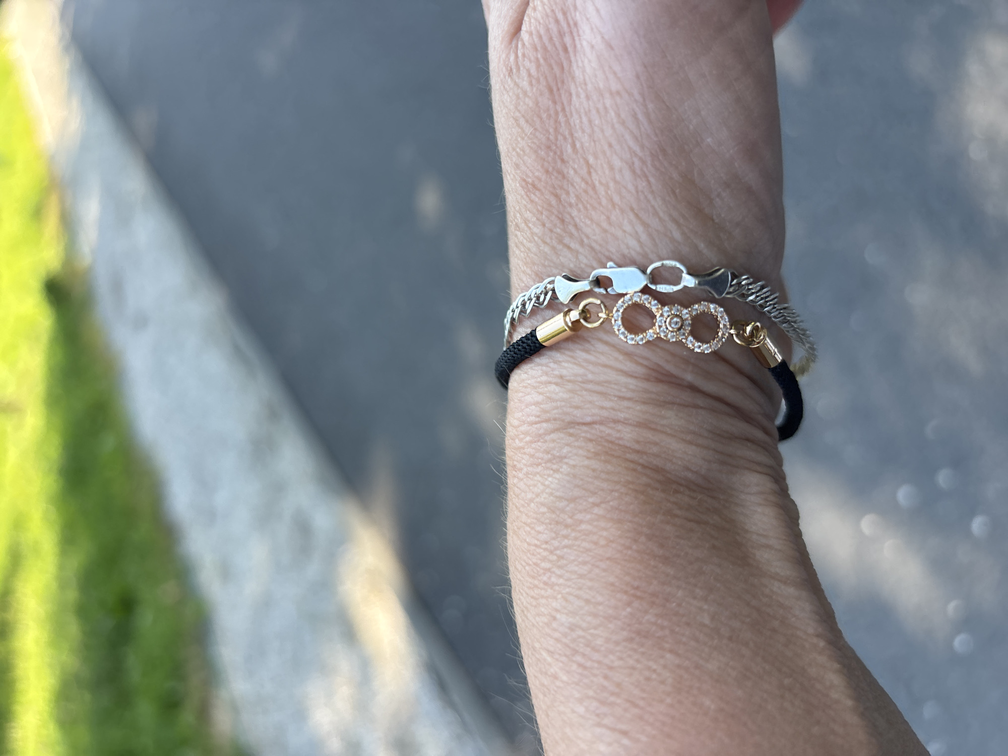 In an outdoor environment, a person's wrist is shown wearing two bracelets. One bracelet is black and features a gold clasp with a dangling charm. This charm is circular and encrusted with small, clear stones. The second bracelet is silver and has a more intricate, chain-like design. Both bracelets are positioned on the wrist, with the black bracelet appearing to be worn underneath the silver one. The background of the image is blurred, showing hints of green and grey, possibly indicating a natural or urban setting with greenery and a paved surface.