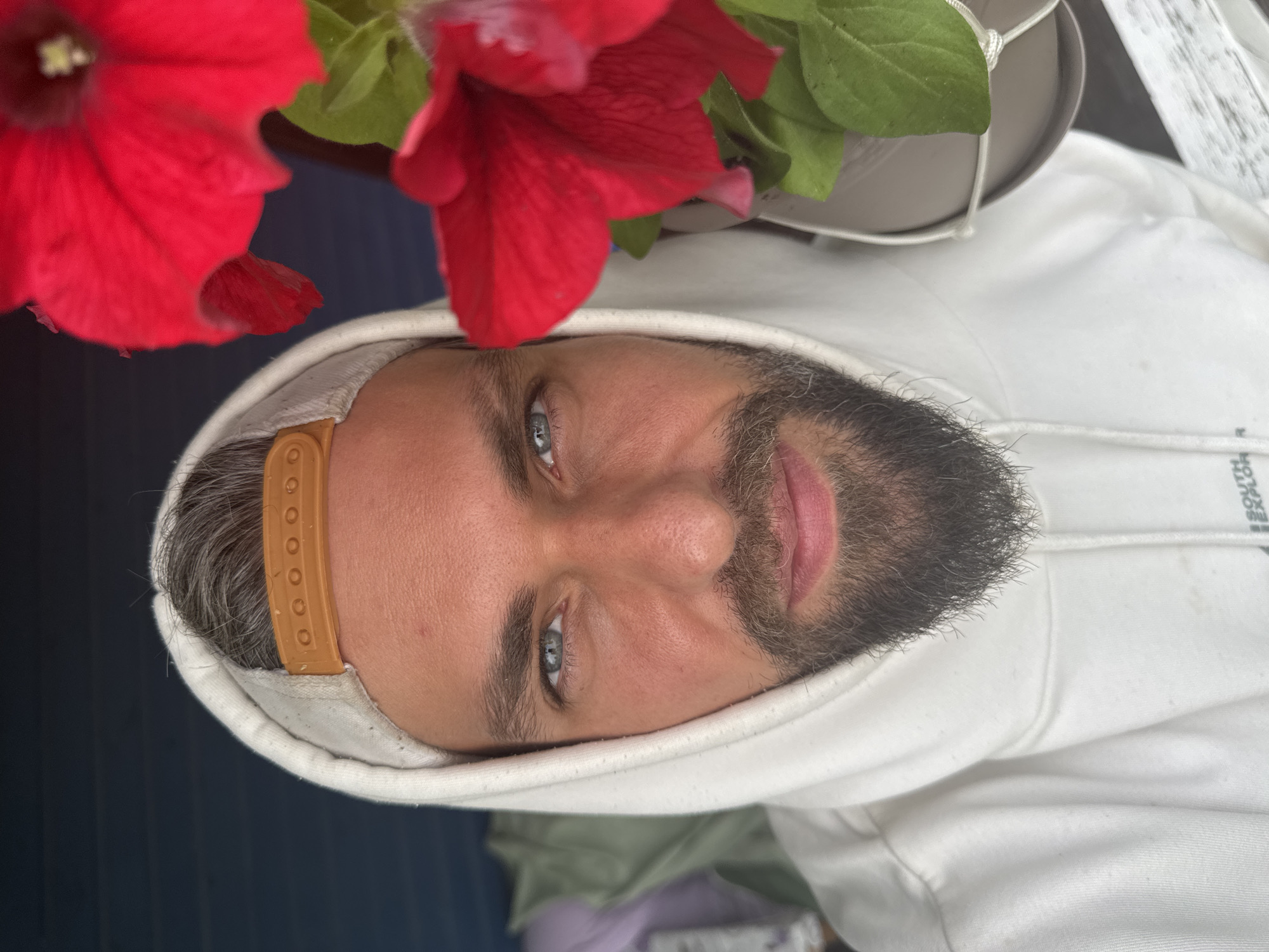 A man wearing a white hoodie and a tan baseball cap is shown in a close-up portrait. He has blue eyes, a beard, and grey hair. The background is a dark blue wall and red flowers are in the foreground. The image does not provide any information about the location in Kuti, Russia.