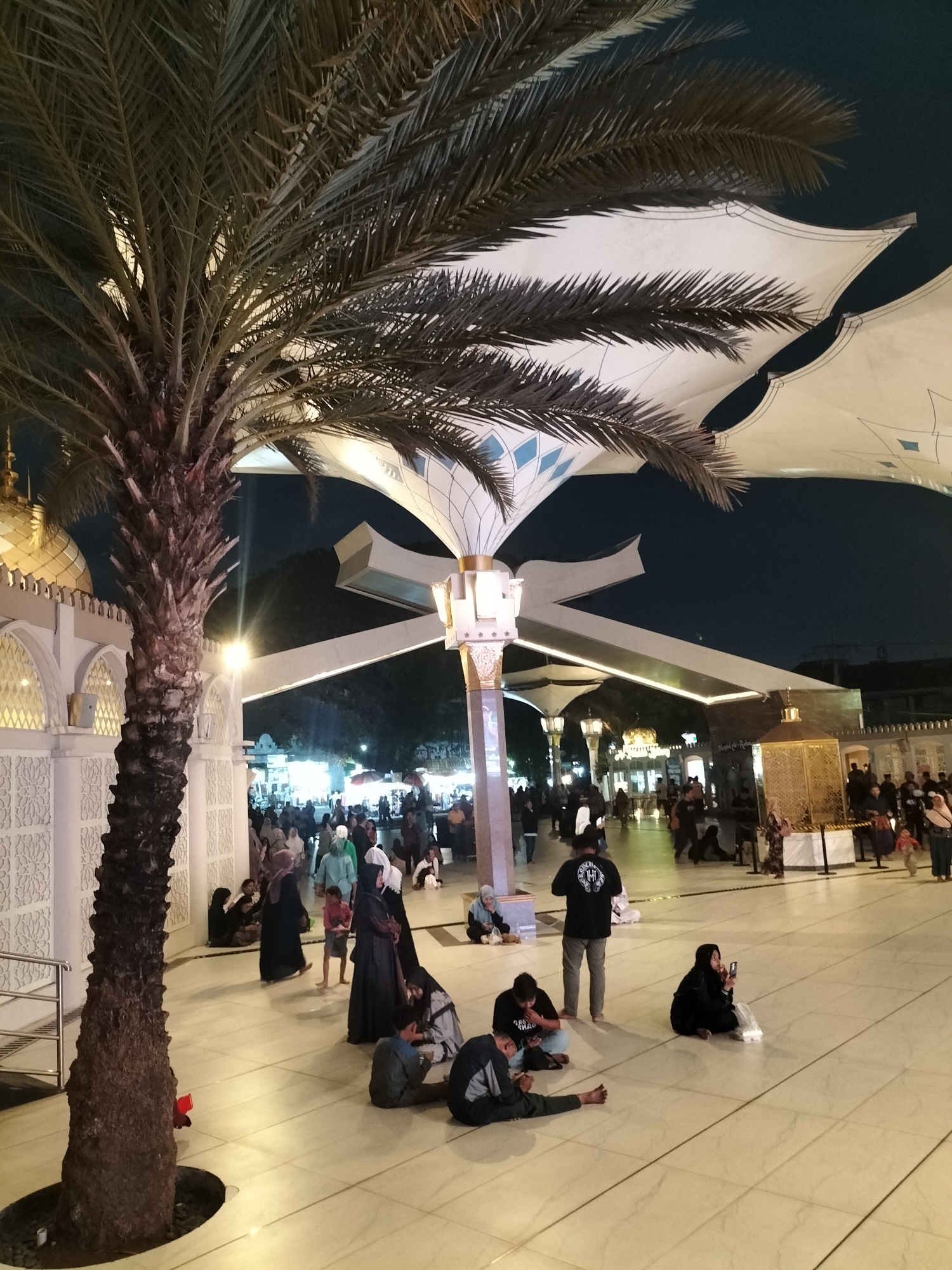 The image depicts an outdoor courtyard at night, likely part of a religious site in Blitar, Indonesia. A large palm tree stands prominently in the foreground on the left, its trunk textured and its fronds casting shadows. Behind the tree, a building with white walls and ornate latticework is visible, topped by a golden dome. Large, white, tensile fabric canopies with decorative blue diamond patterns are suspended overhead, providing shade.
In the courtyard, numerous people are present. Several individuals are sitting on the tiled floor, including a group of men in casual clothing and a woman in a black hijab holding a phone. Other people are standing and walking, some wearing religious attire such as hijabs and robes. In the background, a bustling area with stalls and bright lights suggests a marketplace or food court. The overall scene conveys a sense of community gathering and nighttime activity at a cultural or religious landmark in Blitar.