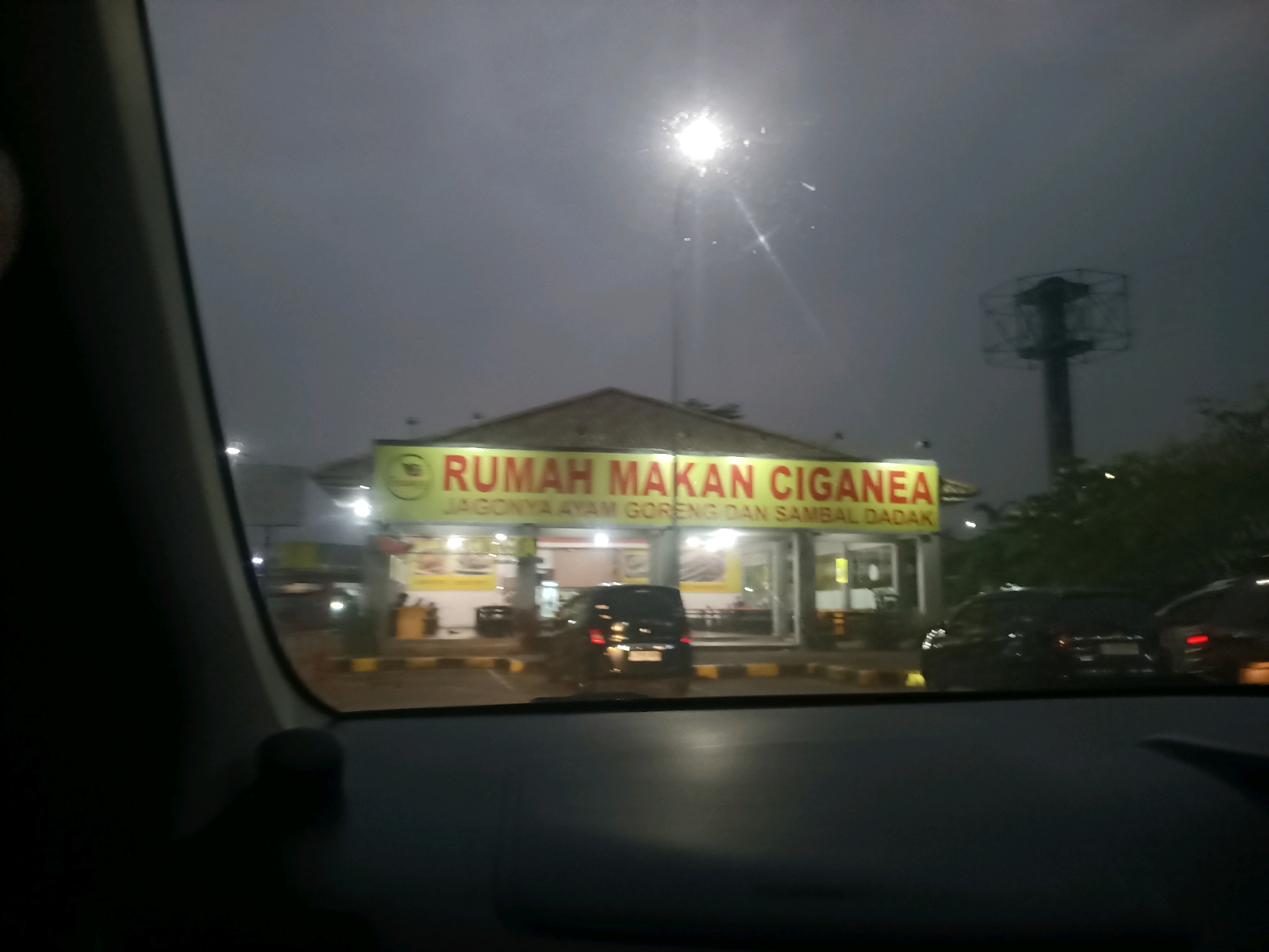 The image shows a view from inside a vehicle, looking out at a restaurant called "RUMAH MAKAN CIGANEA" which specializes in "JAGONNYA AYAM GORENG DAN SAMBAL DADAK". The restaurant sign is illuminated in yellow with red lettering. In front of the restaurant, there are several cars parked. The scene appears to be at dusk or night, as evidenced by the low light conditions and the artificial lighting from the restaurant and streetlights. In the background, there is a tall, dark structure that resembles a radio tower or a pylon. The weather appears overcast. The car the photo is taken from has a dashboard and part of the windshield frame visible in the foreground. The overall impression is that of a roadside eatery in Indonesia, likely in the city of Karawang, as suggested by the restaurant's name, which is a well-known local brand there.