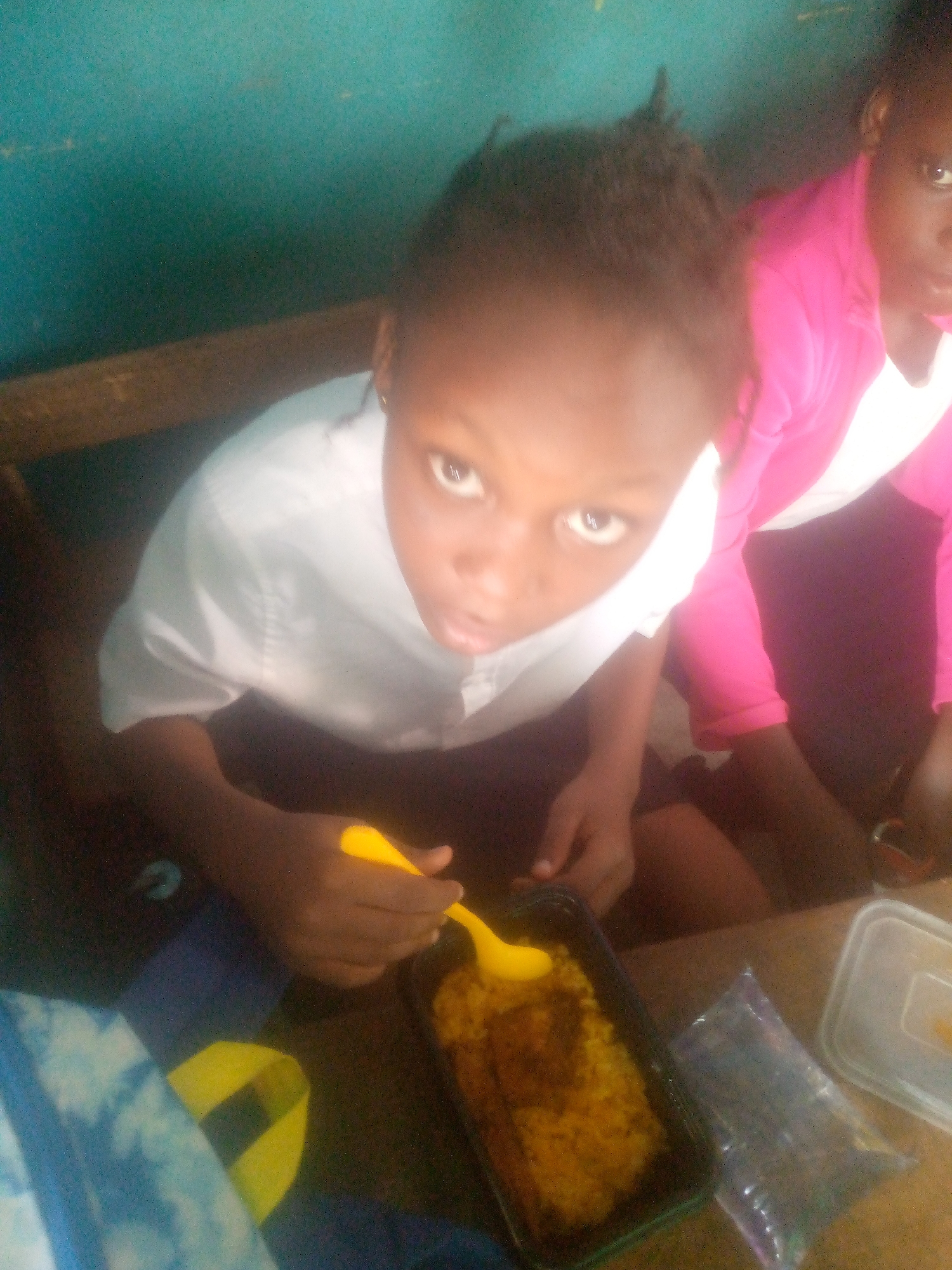 A high-angle shot shows a young person with dark skin eating food with a yellow spoon from a black rectangular container. A second person, partially visible, is wearing a pink top. The setting appears to be indoors, possibly a school or similar institution, though the specific location in the city of Unknown, Unknown is unclear from the image. There is a blue backpack and clear plastic containers on the table.