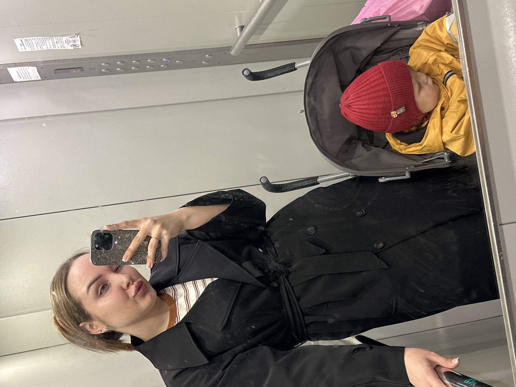 A woman takes a selfie in a mirrored elevator with a baby in a stroller. The woman has light brown hair and is wearing a black trench coat. The baby is wearing a red knit hat and a yellow jacket. A pink bag is in the stroller. The elevator's control panel and indicator lights are visible. The scene appears to be indoors. The image was taken in Chaikovsky, Russia.