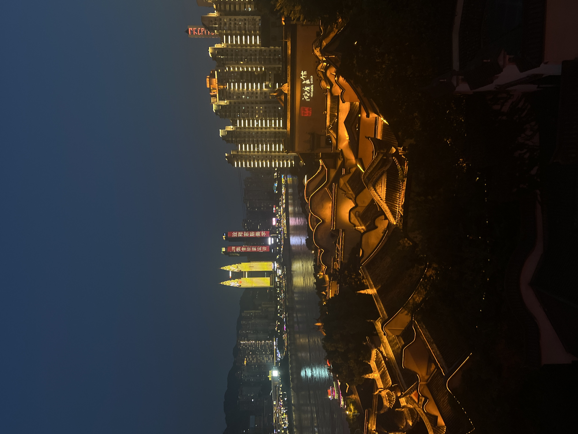 This is a nighttime aerial shot of a waterfront cityscape in Yuzhong District, China. The foreground features a collection of traditional Chinese-style buildings with illuminated, curved roofs and intricate architectural details, outlined by warm yellow lights. Beyond these, a wide river reflects the lights of the city. On the far bank, a modern skyline of tall buildings is visible, many of which are lit with white and yellow lights. Two prominent, bright yellow twin towers stand out in the center of the modern skyline, and a building with Chinese characters illuminated in red and white is also visible. The sky is a dark, deep blue, indicating nighttime. No people are discernible in the image.