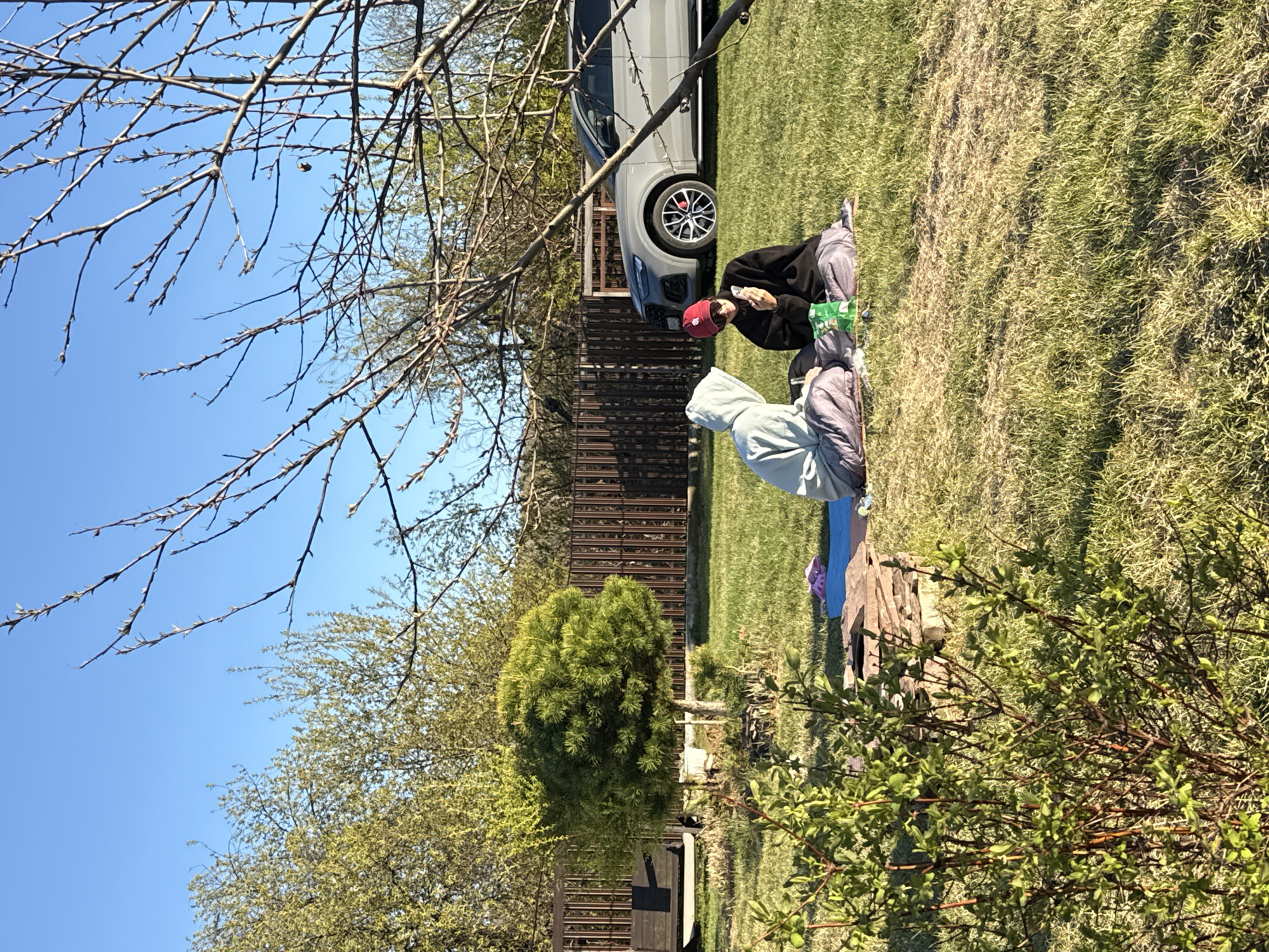 The image shows a daytime outdoor scene in what appears to be a backyard or grassy area. Two individuals are sitting on the grass. One is wearing a dark jacket and a red cap, appearing to be holding a cell phone. Next to this individual is another person with a grey hoodie. Behind the people is a dark-colored SUV parked in front of a brown wooden fence. A small evergreen tree and a deciduous tree are in the background. The sky is a clear blue, partially obscured by bare tree branches. Given the presence of vegetation and residential characteristics, it’s plausibly located in a suburban area. Since the prompt requested that the image be located in Emel'yanovo, Russia, this is assumed to be that location.